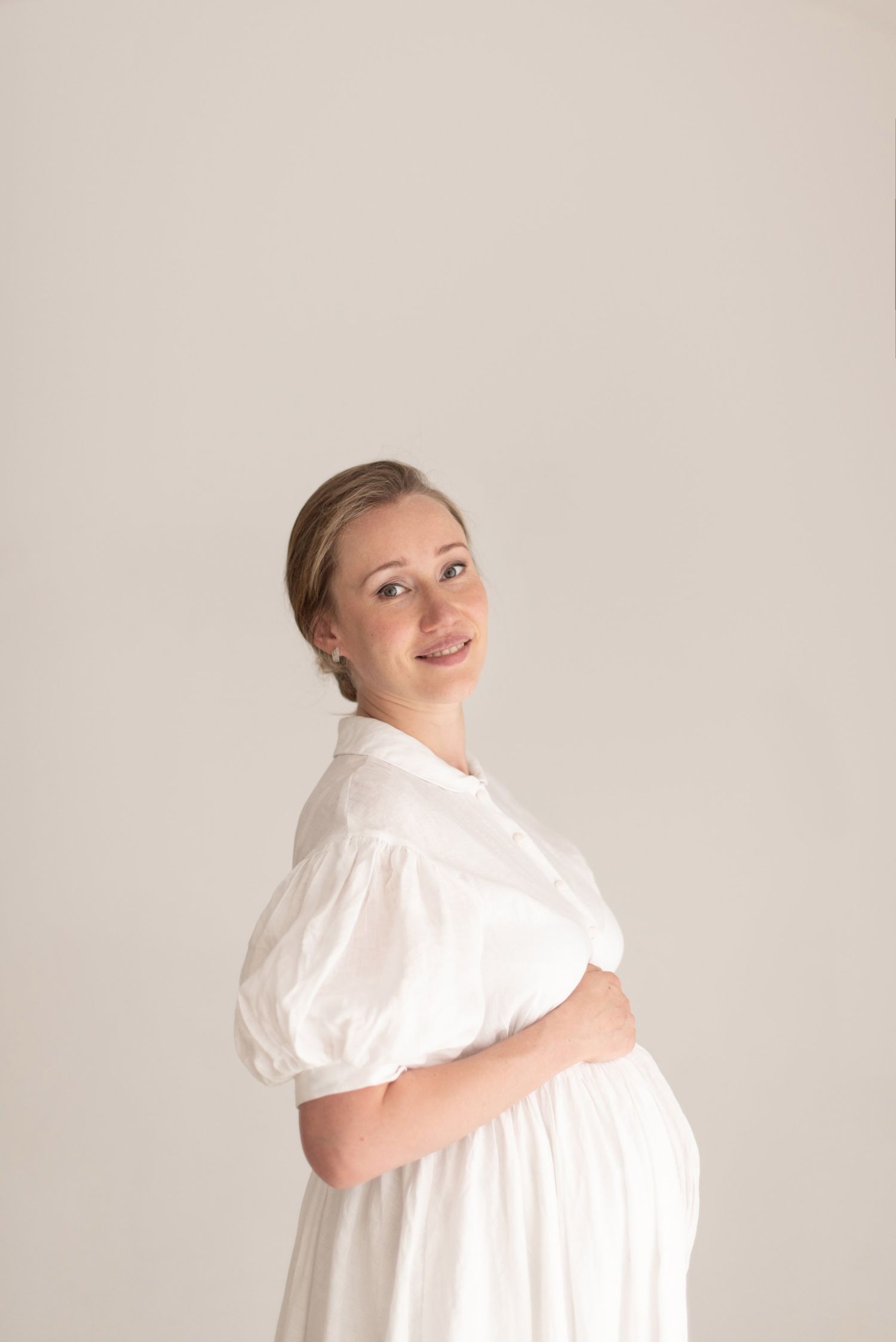 Side profile of pregnant woman in flowing white dress against neutral background.