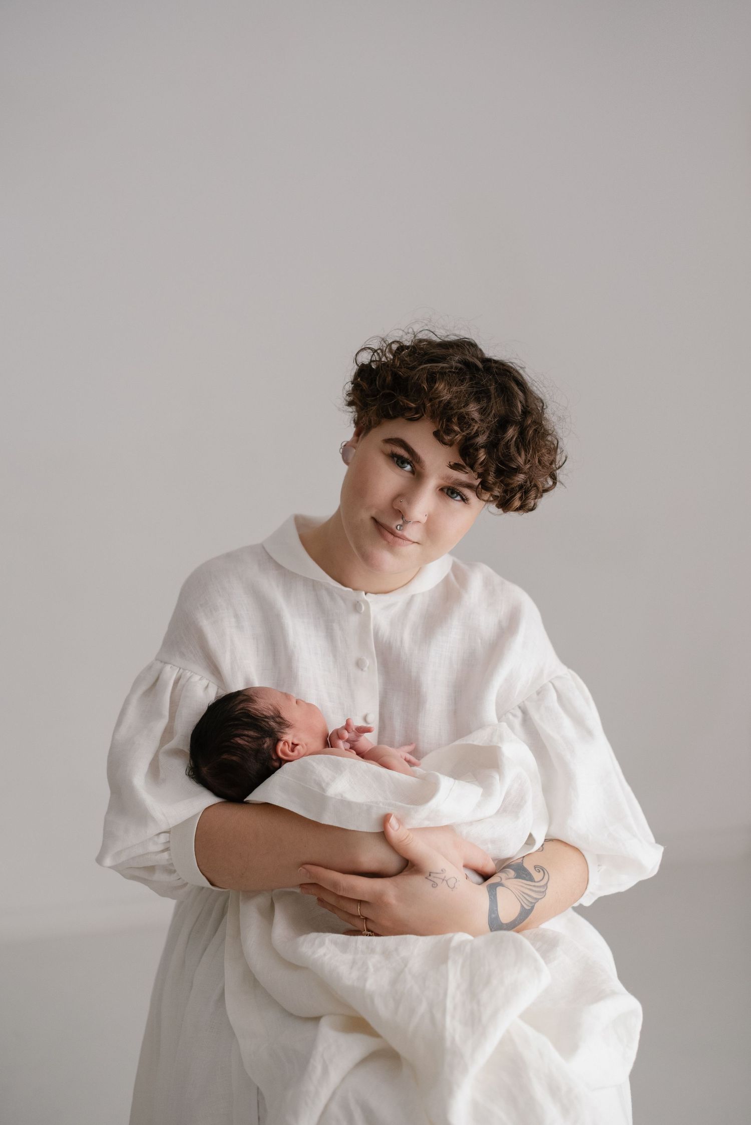 Parent in white linen dress holding newborn baby against neutral background in minimalist studio portrait.