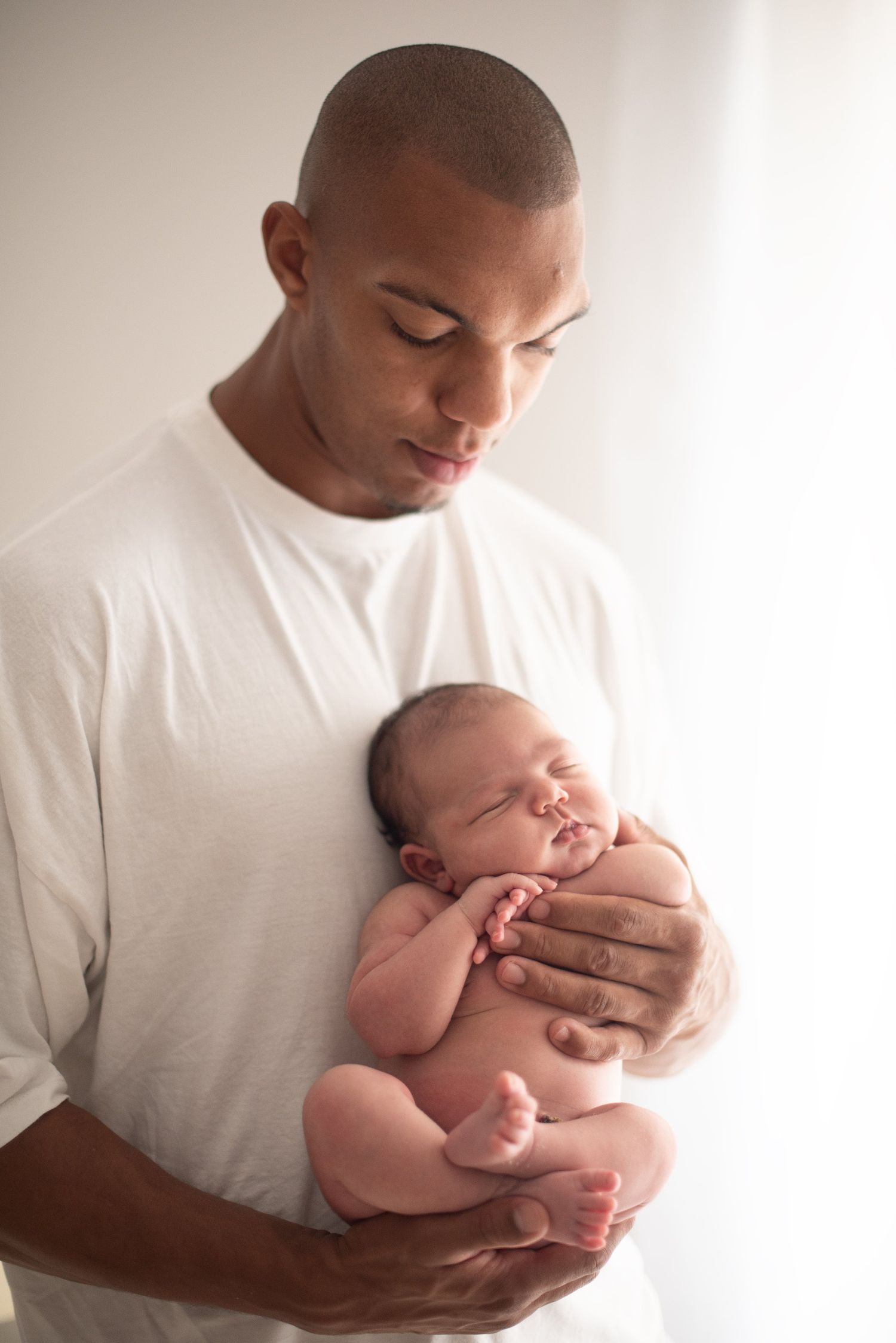 Gentle father in white shirt holding sleeping newborn baby by window in tender moment.