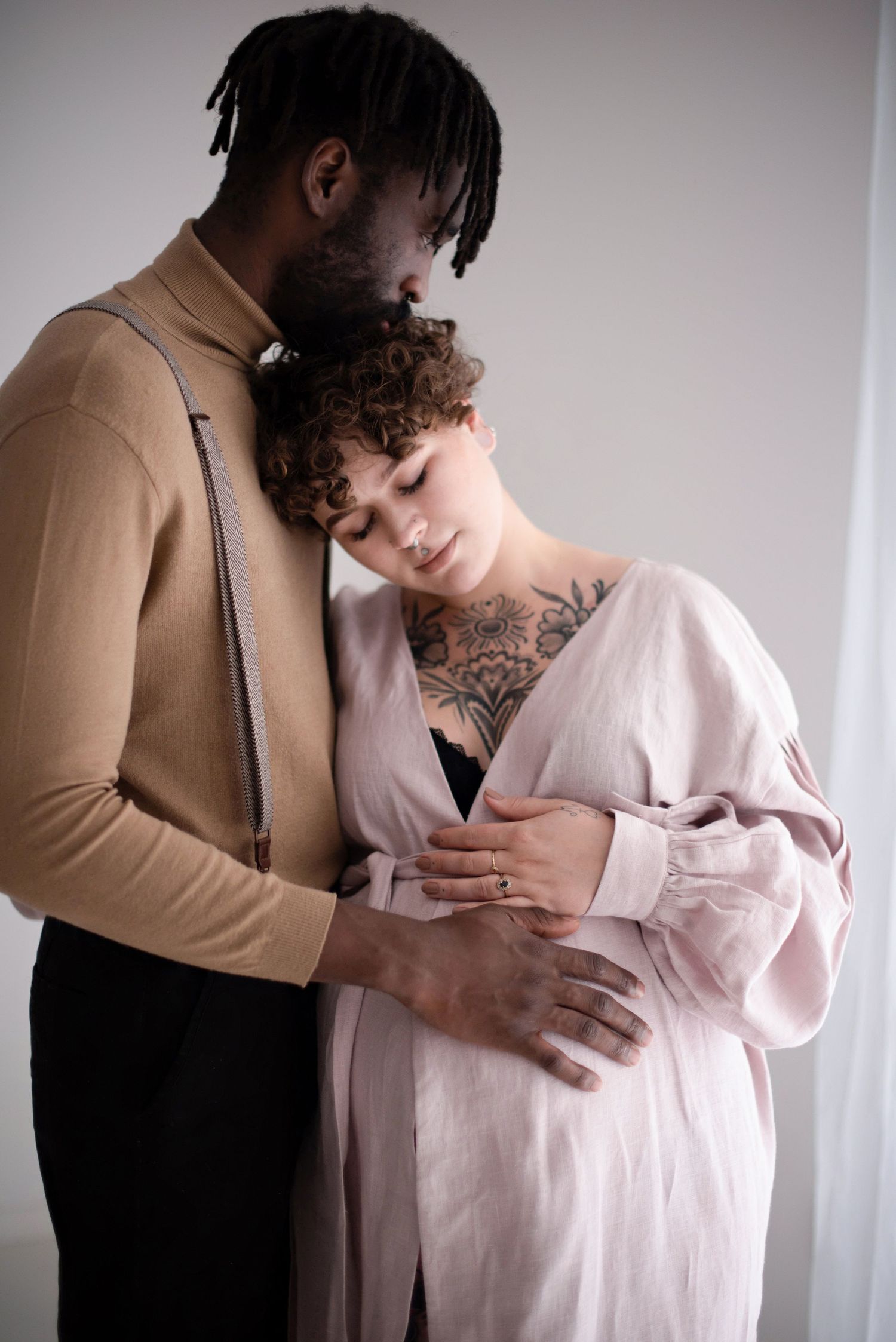 Intimate maternity photo of couple embracing, person wearing tan sweater holding partner in pink robe against white wall.