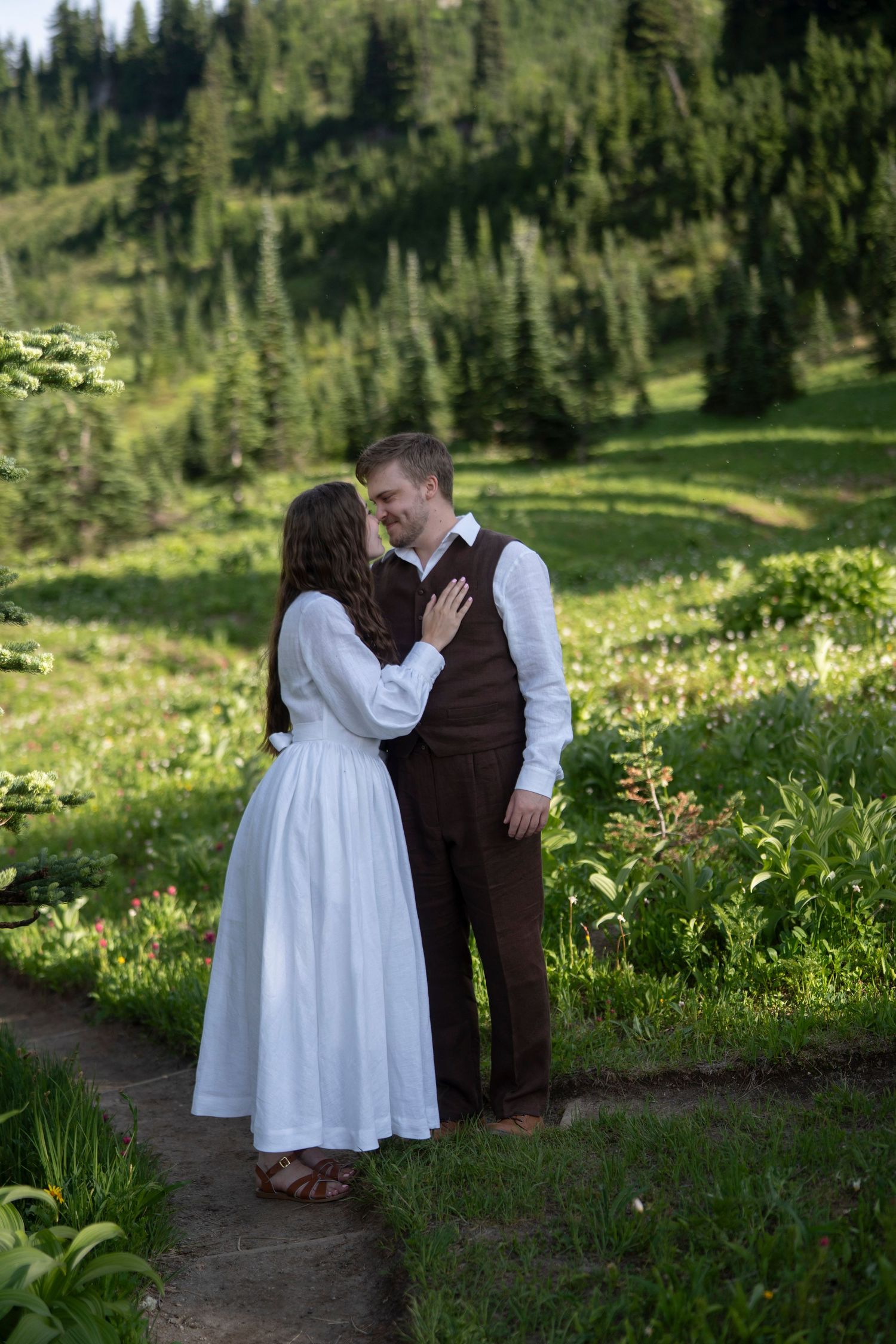 A couple shares a romantic moment on a mountain trail surrounded by wildflowers and evergreen trees in summer.