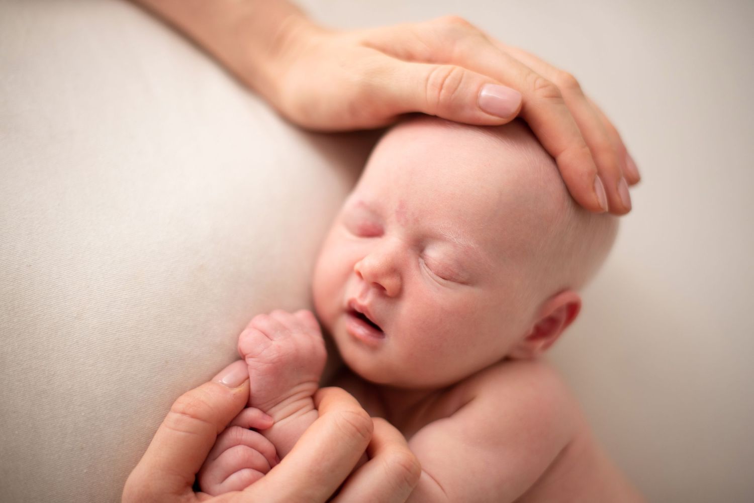 A sleeping newborn baby with light skin tone is gently cradled by caring hands against a neutral background.