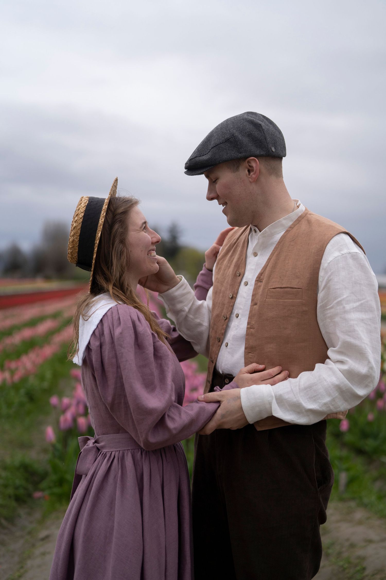 A romantic vintage-style couple embraces in period clothing while standing among blooming tulip fields on an overcast day.