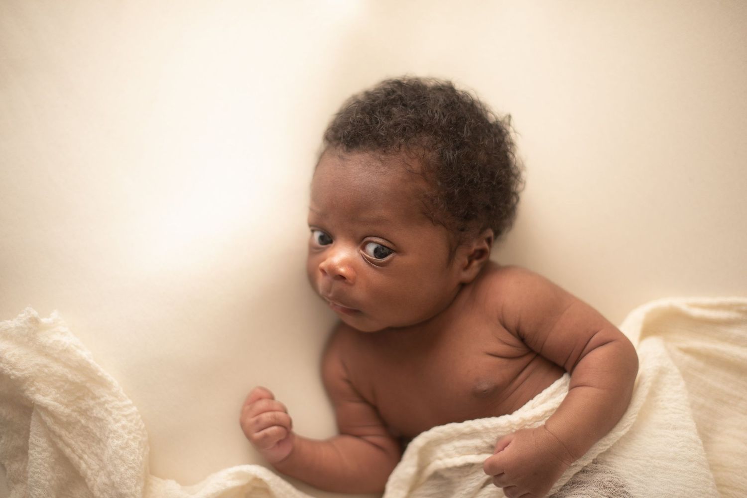 Alert newborn baby resting on cream colored fabric in studio portrait.