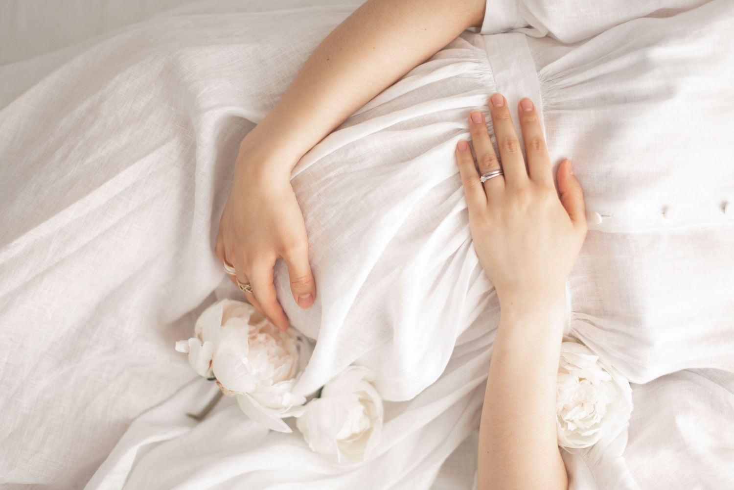 Delicate hands rest on soft white fabric in ethereal maternity photo composition.