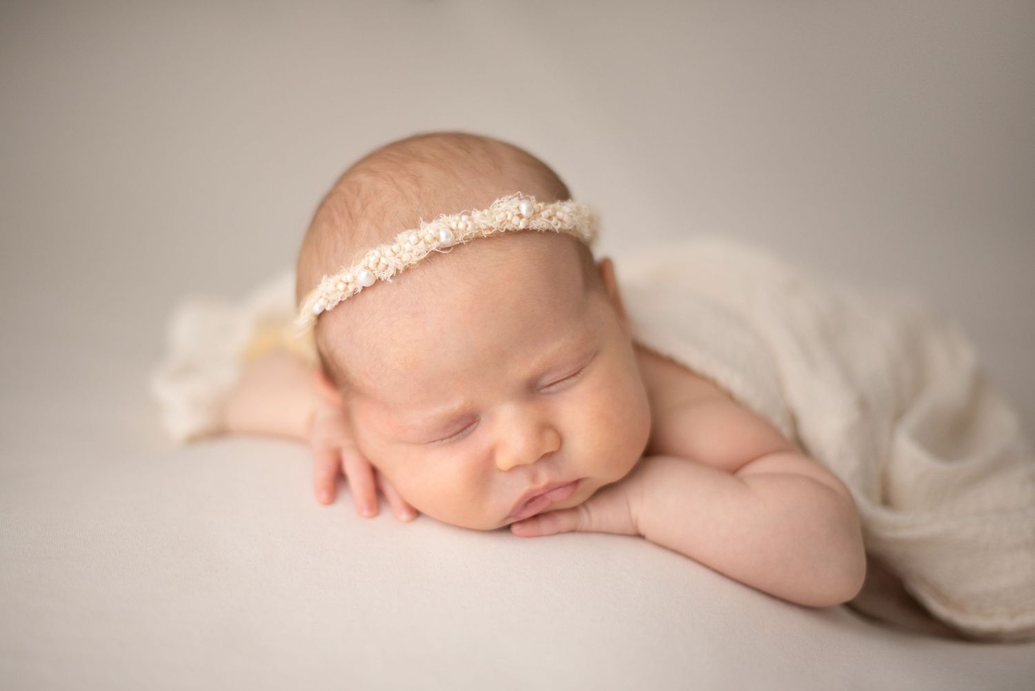 Peaceful newborn posed on white backdrop wearing delicate beige headband during professional photo shoot.