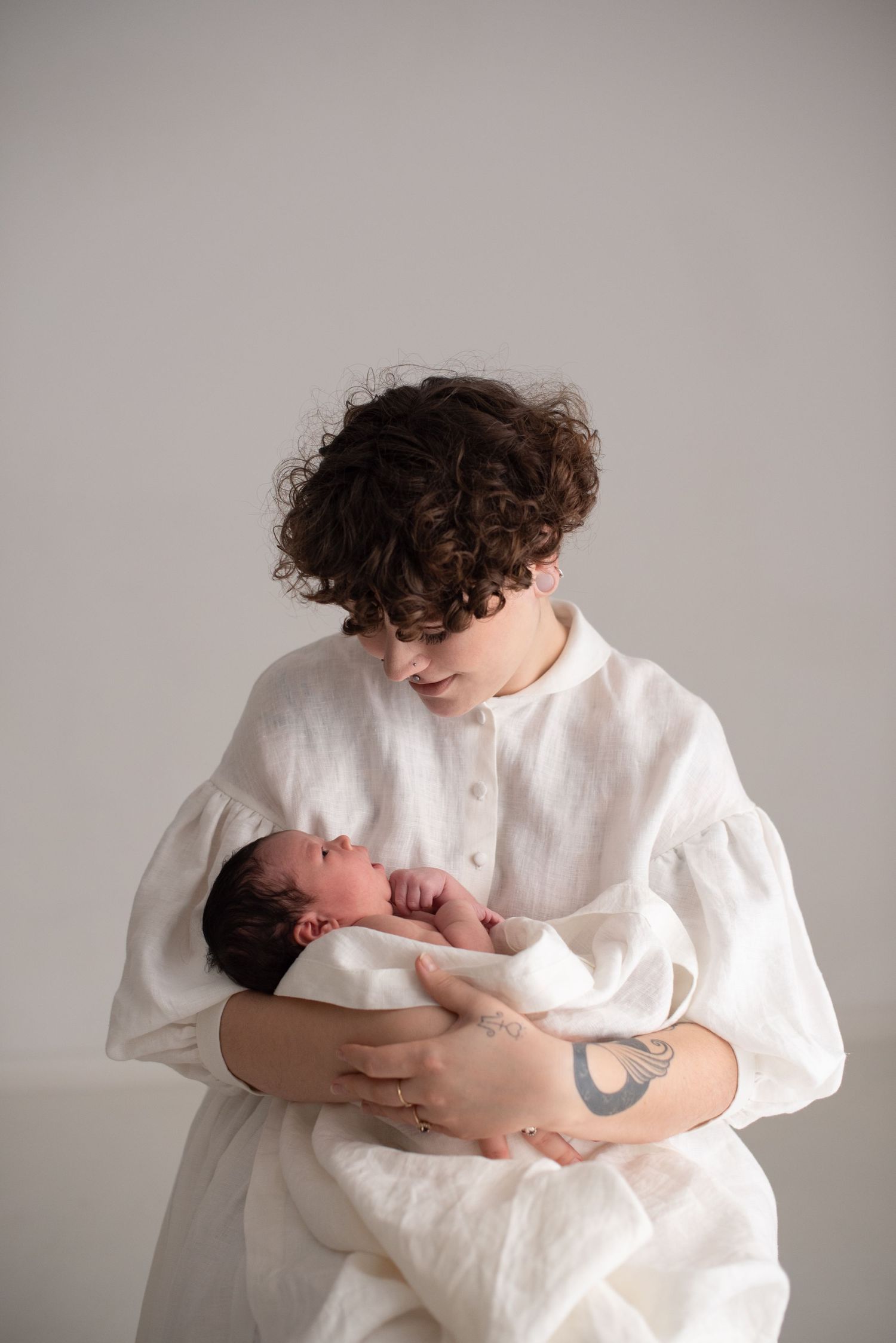 Parent in billowing white dress gazes lovingly at newborn baby in arms against neutral backdrop.