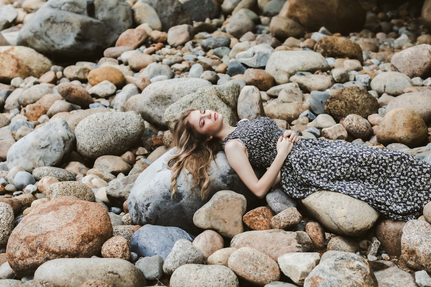 Rachel Delong, musician, lays on river rocks wearing a long gray floral dress for a portrait shoot in the White Mountains North Conway NH.