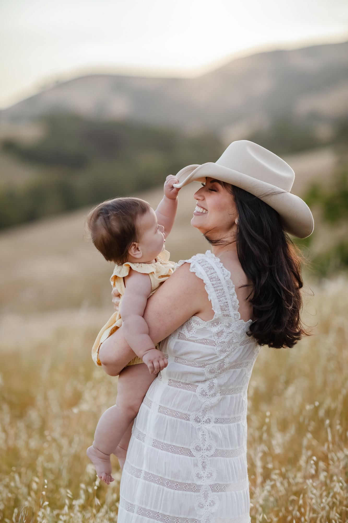 Baby girl wearing pale yellow pulls down dark haired mother's cream colored cowboy hat wearing a cream lace dress in golden rolling hills.