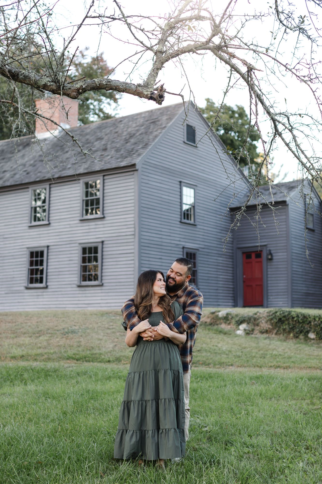 Fiancee embraces girlfriend wearing a green dress from behind in front of the Nathan Meriam House in Concord, MA for engagement photos.
