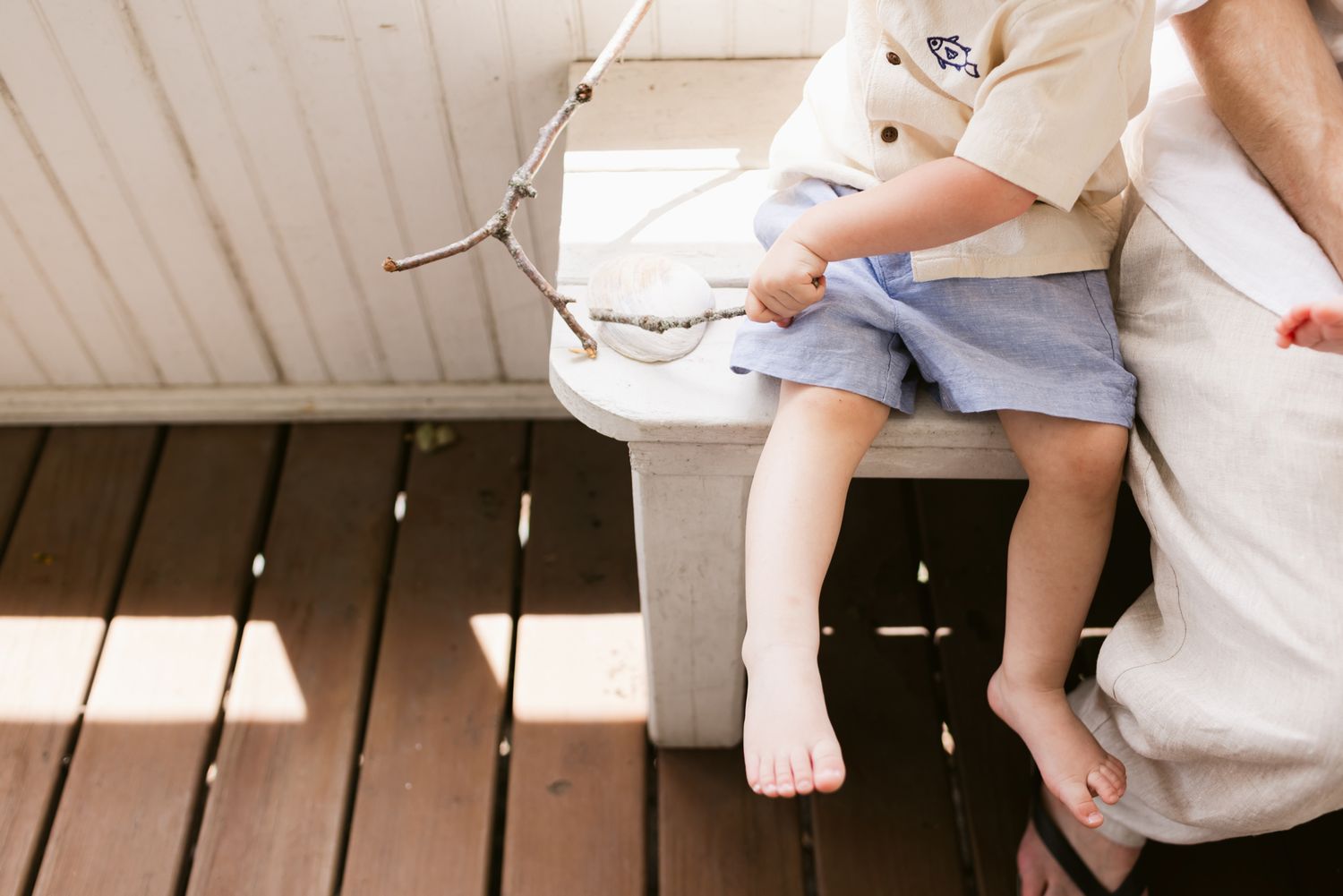 Toddler boy wears blue shorts and cream short sleeves plays with a stick on a sunny porch while sitting on a white bench for family photos.