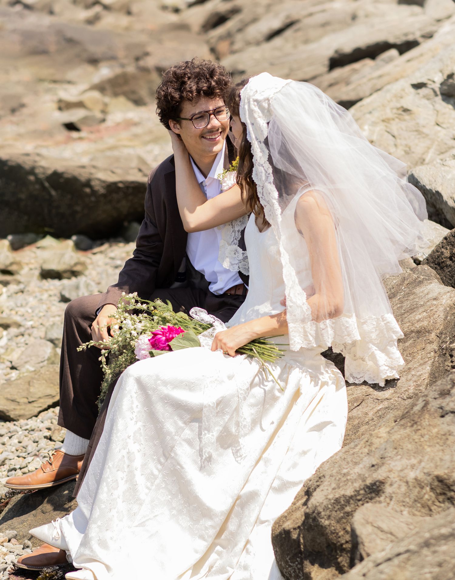 Bride holds grooms face with veil flowing in wind sitting on coastal rocks on a sunny wedding day. Newport County RI wedding photographer