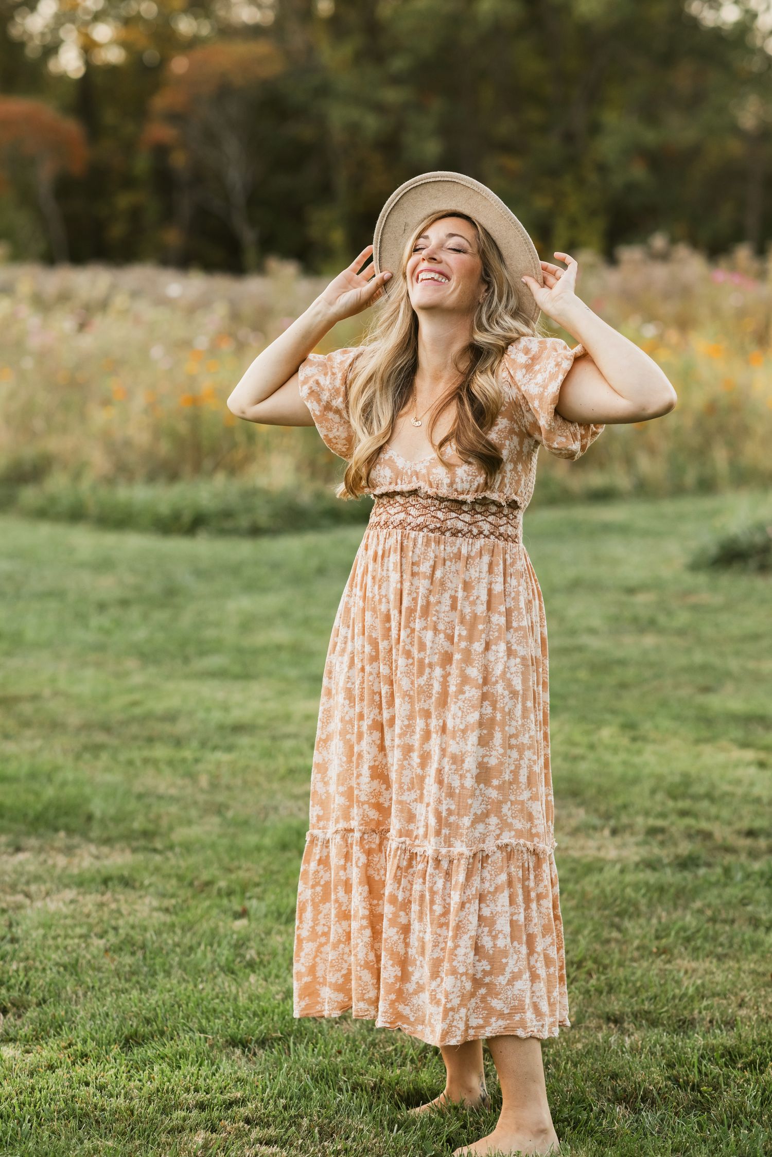 A smiling mother with long blonde hair wearing an orange floral peasant dress and a tan suede hat tilts her head back for a motherhood photo