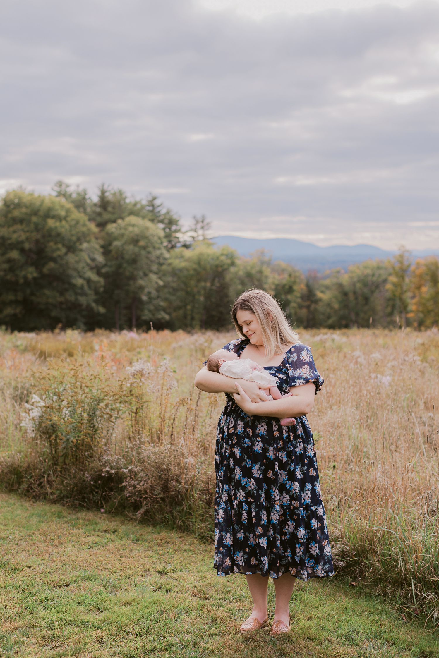 A mother with short blond hair wearing a navy floral dress holds her newborn baby girl in a cream outfit by a golden field in Tamworth, NH.
