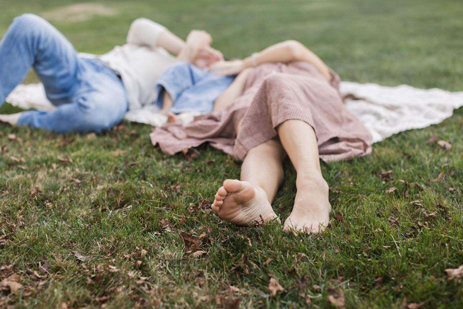 A family lays on a cream crochet blanket on lush green grass with a photographer's focus on the mom's feet for family photos in Stratham, NH