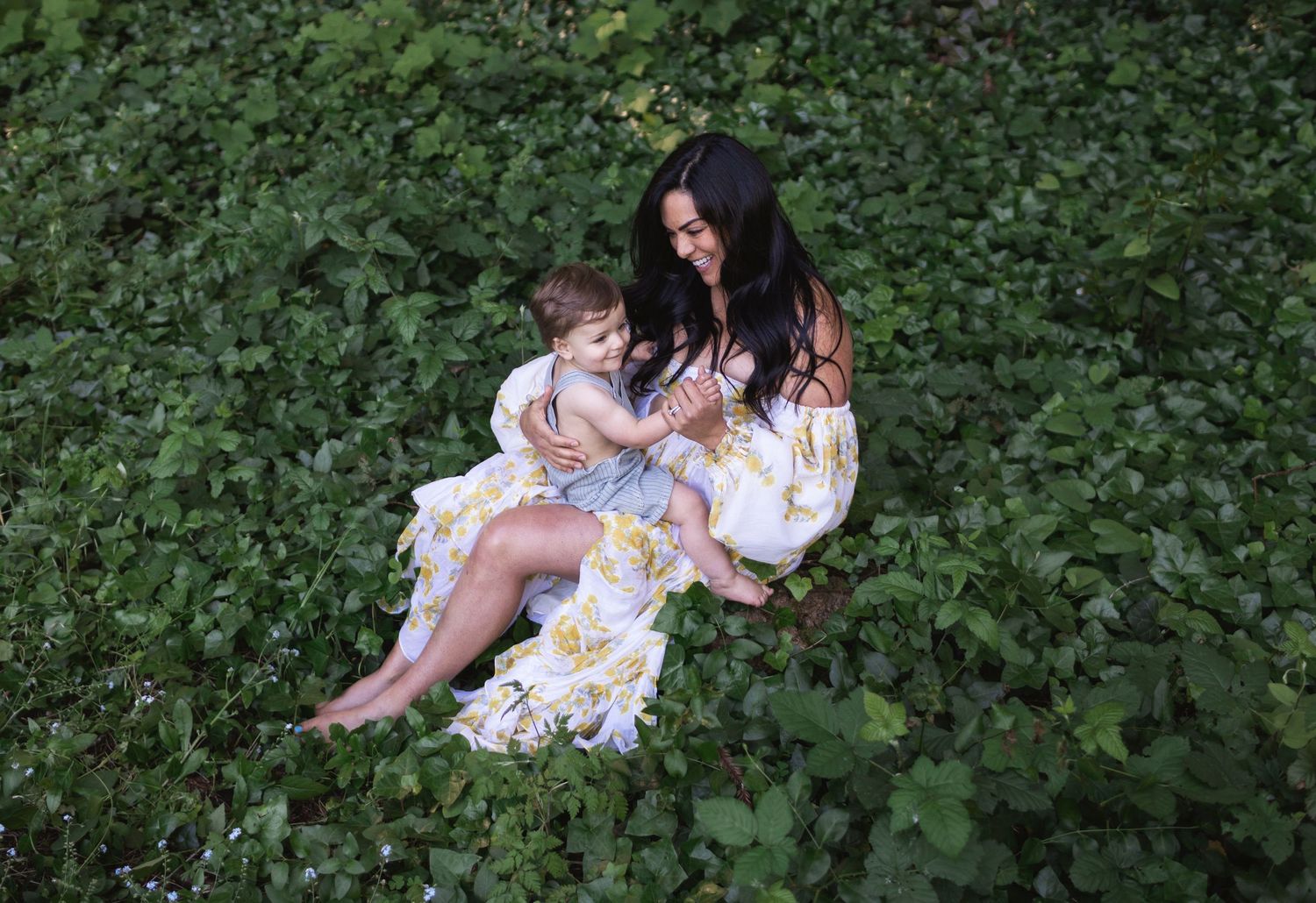 A mother and child sit together among lush green foliage in a garden for a motherhood photo shoot .