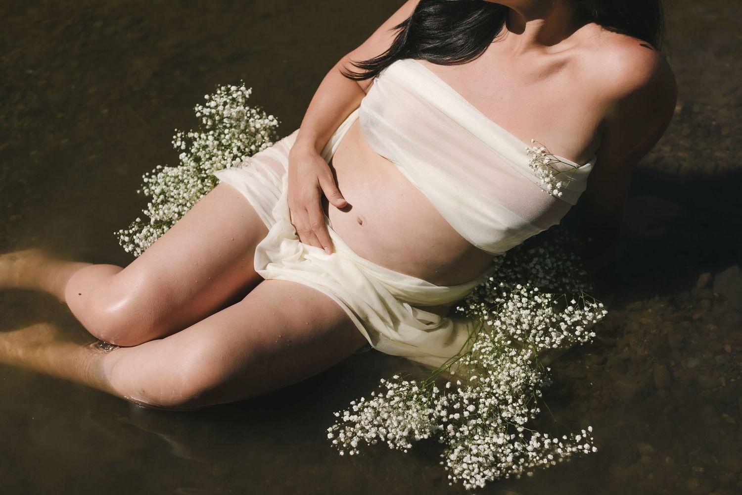 Expectant mother wears a cream mesh covering for maternity photos laying in a shallow river. North Conway Laconia maternity photographer