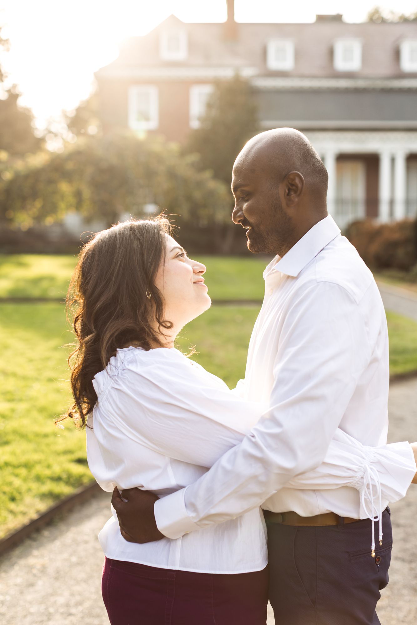 Couple wears white shirts gazes at each other with beautiful sun light upon them in front of a colonial brick building at Garden at Elm Bank