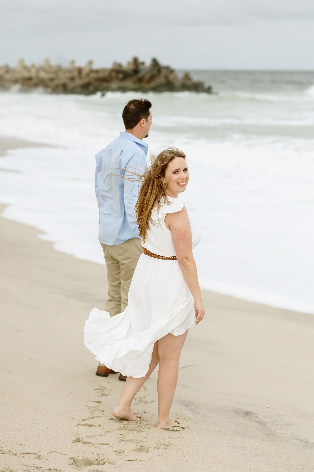 Blond woman in flowy white dress holds hand of man on windy day beach day for engagement photos. Acadia engagement photographer.