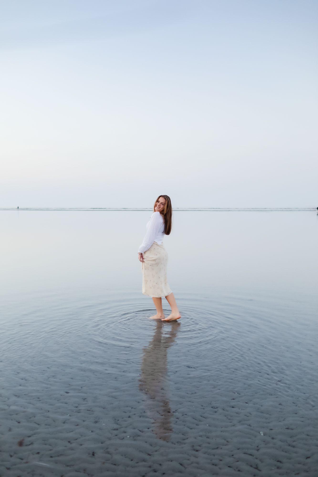 Blond senior girl holds her floral dress posing in shallow water at Wingaersheek Beach in Gloucester, Massachusetts for senior portraits.