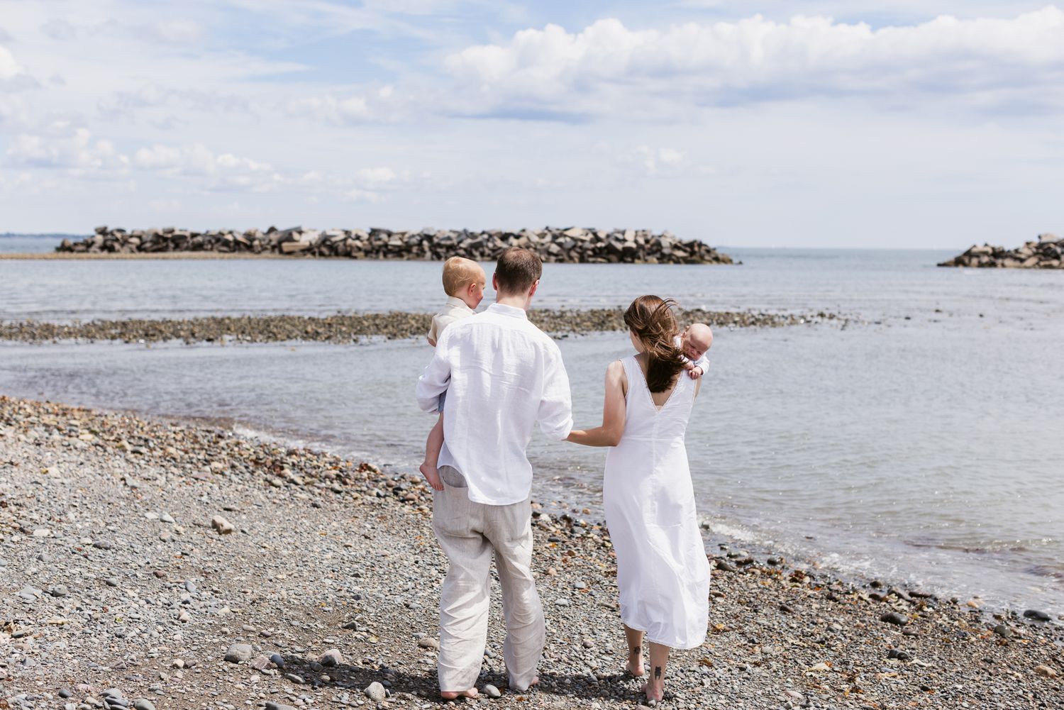 Family wearing white walks backward holding hands on rocky shoreline on a sunny day for newborn family photos. Ipswich newborn photographer.