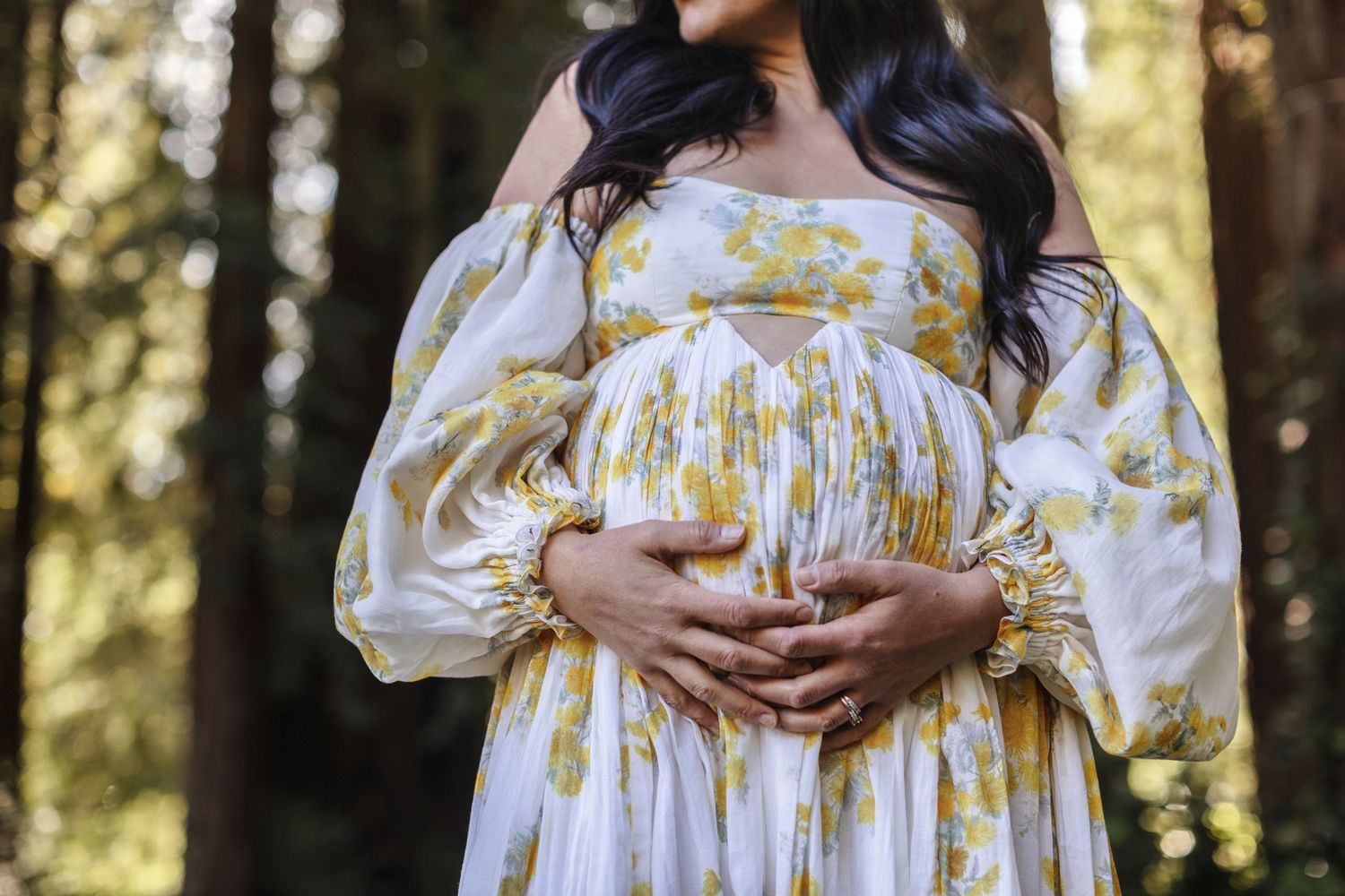 Expectant mother in floral maternity gown embraces baby bump in a forest with bokeh. Concord Lakes Region North Shore maternity photographer