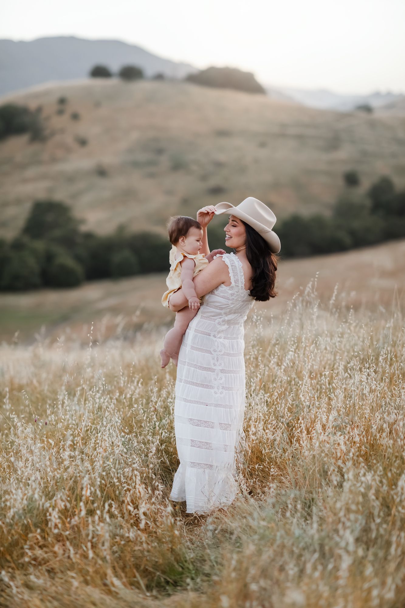 Mother in lace area, dress wearing a cream cowboy hat holds baby girl in yellow dress in open rolling grassy field on cloudy day.