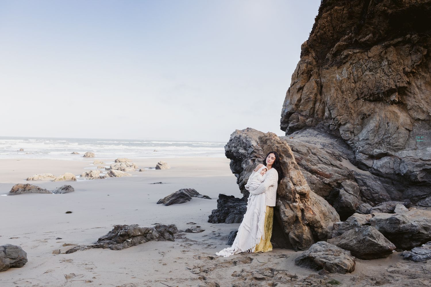 Mother with long dark hair wearing a yellow lace dress and cream sweater holds a baby in a blanket leaning against a large coastal rock.