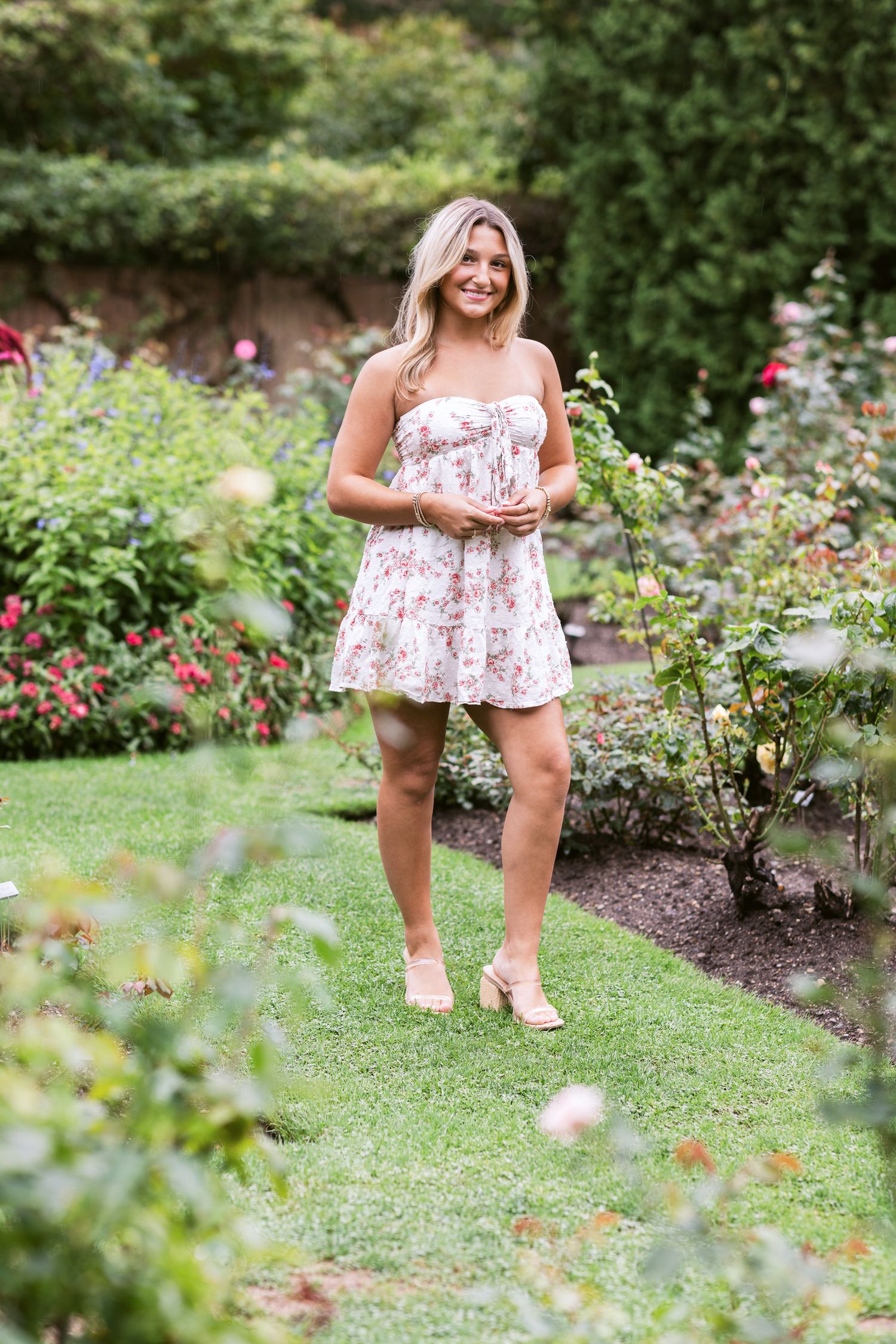 A blond senior girl holds her hands standing in a rose garden for senior photos at Fuller Gardens in North Hampton, New Hampshire.