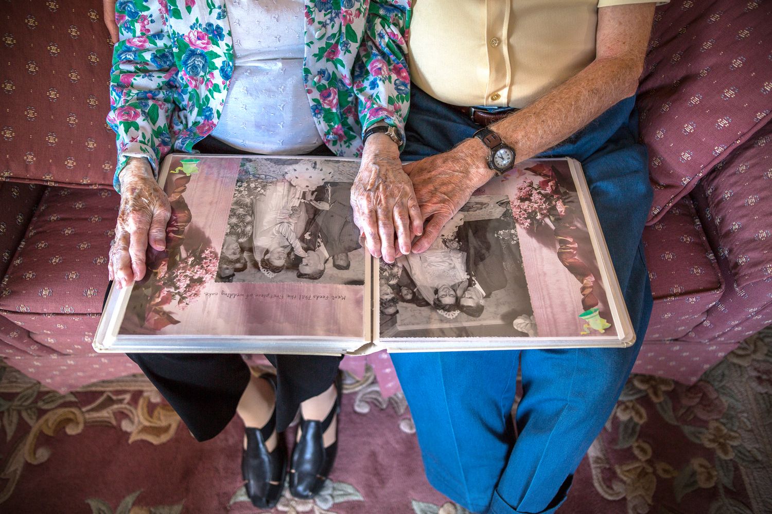 Elderly couple lay their hands on each others over their wedding album while sitting on their mauve floral couch. New Hampshire photographer