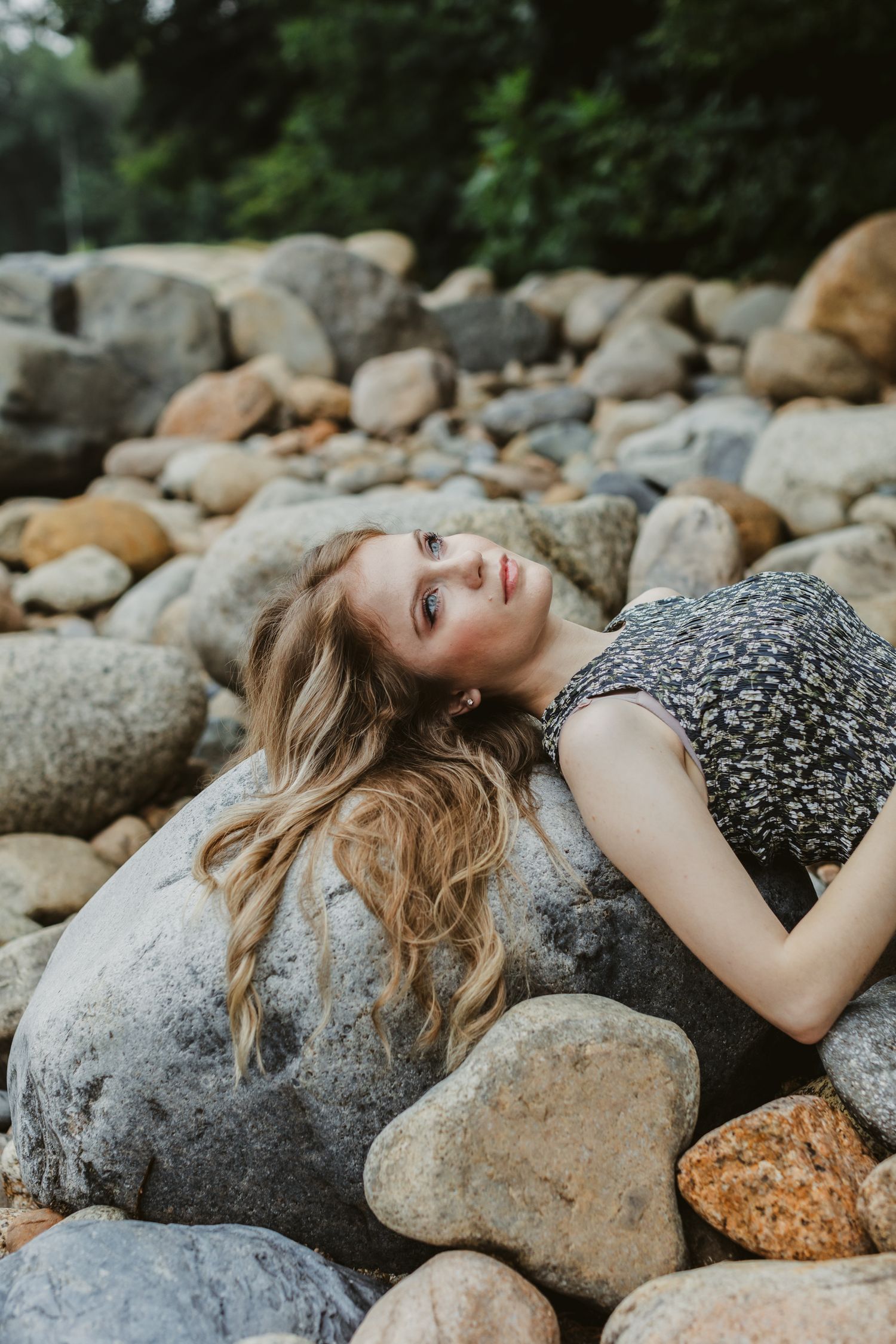 Rachel Delong, Christian musician, wears a gray floral dress and lays her head with long auburn hair on a large river rock in Lincoln, NH.