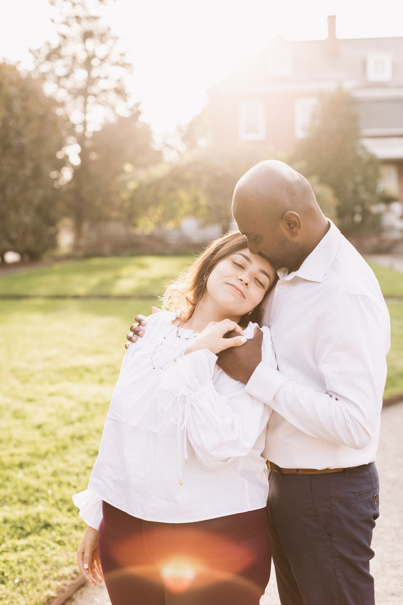 Woman rests head on man's shoulder as her holds her hand with sun flare upon them in front of brick colonial building at Garden at Elm Bank