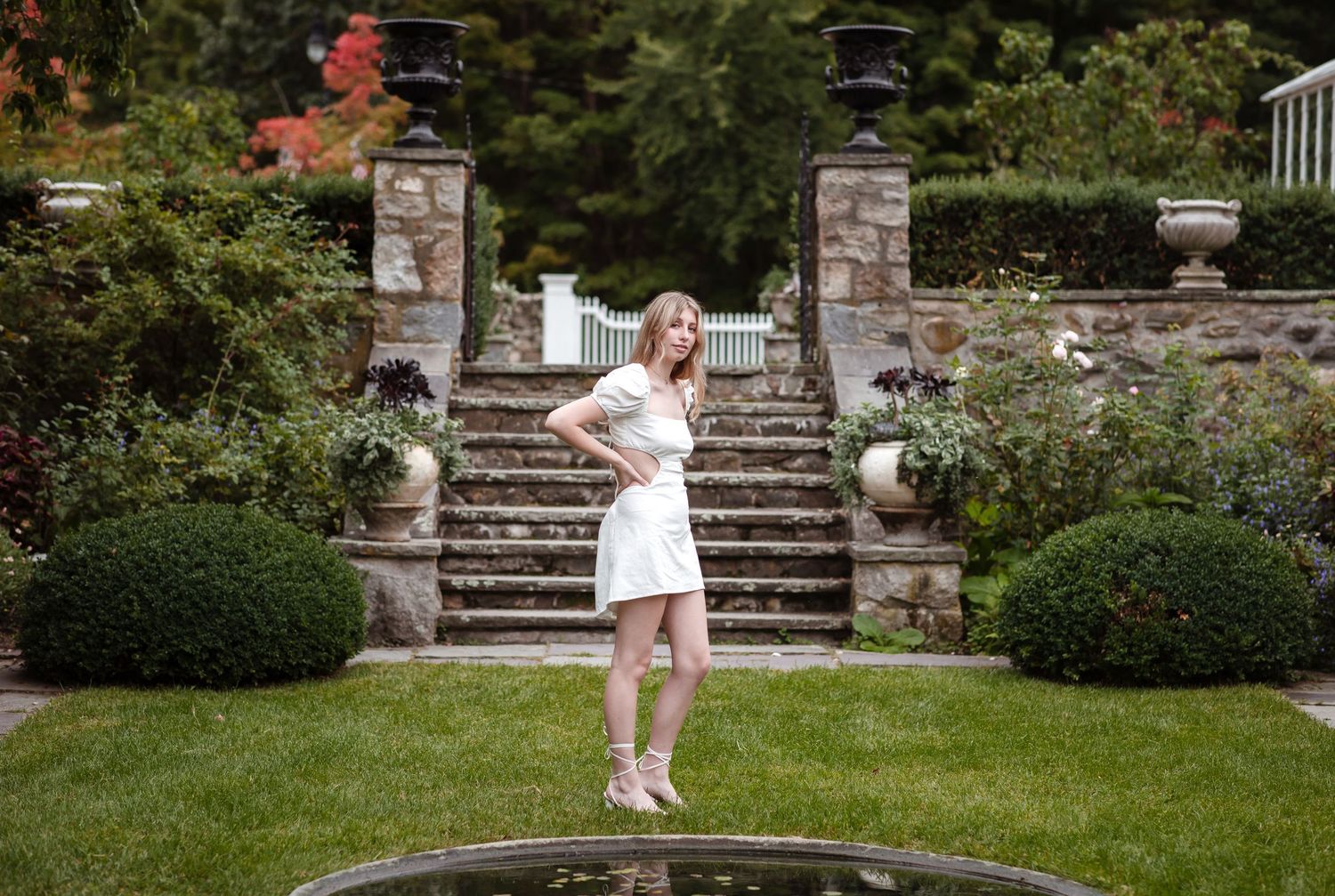 Blond senior girl wears short white dress poses in front of small frog pond in a garden. with stone steps at Steven Coolidge Estate Andover