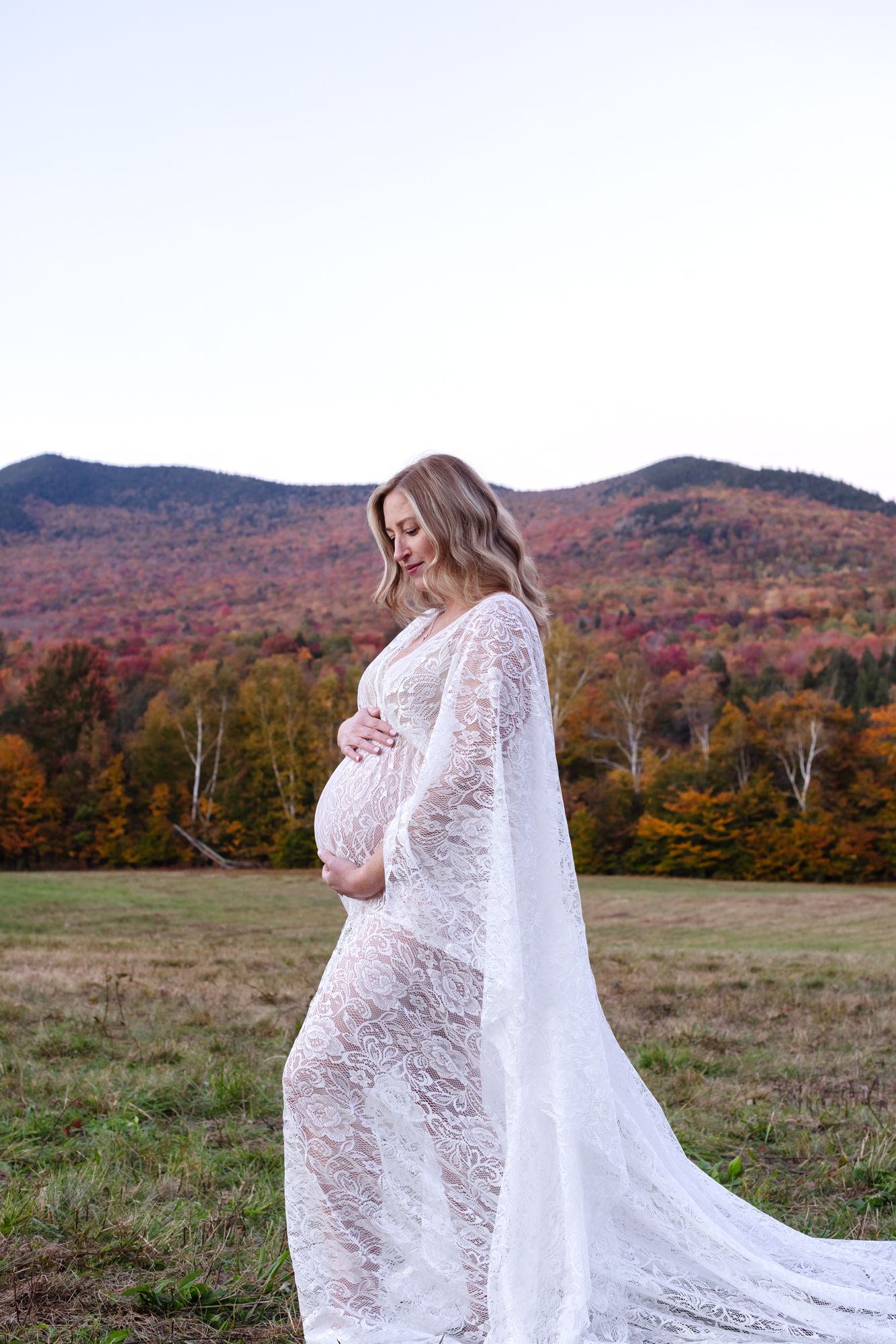 Expectant mother wears a white lace maternity gown posing against a soft vibrant foliage mountain background for maternity photos in Stowe.
