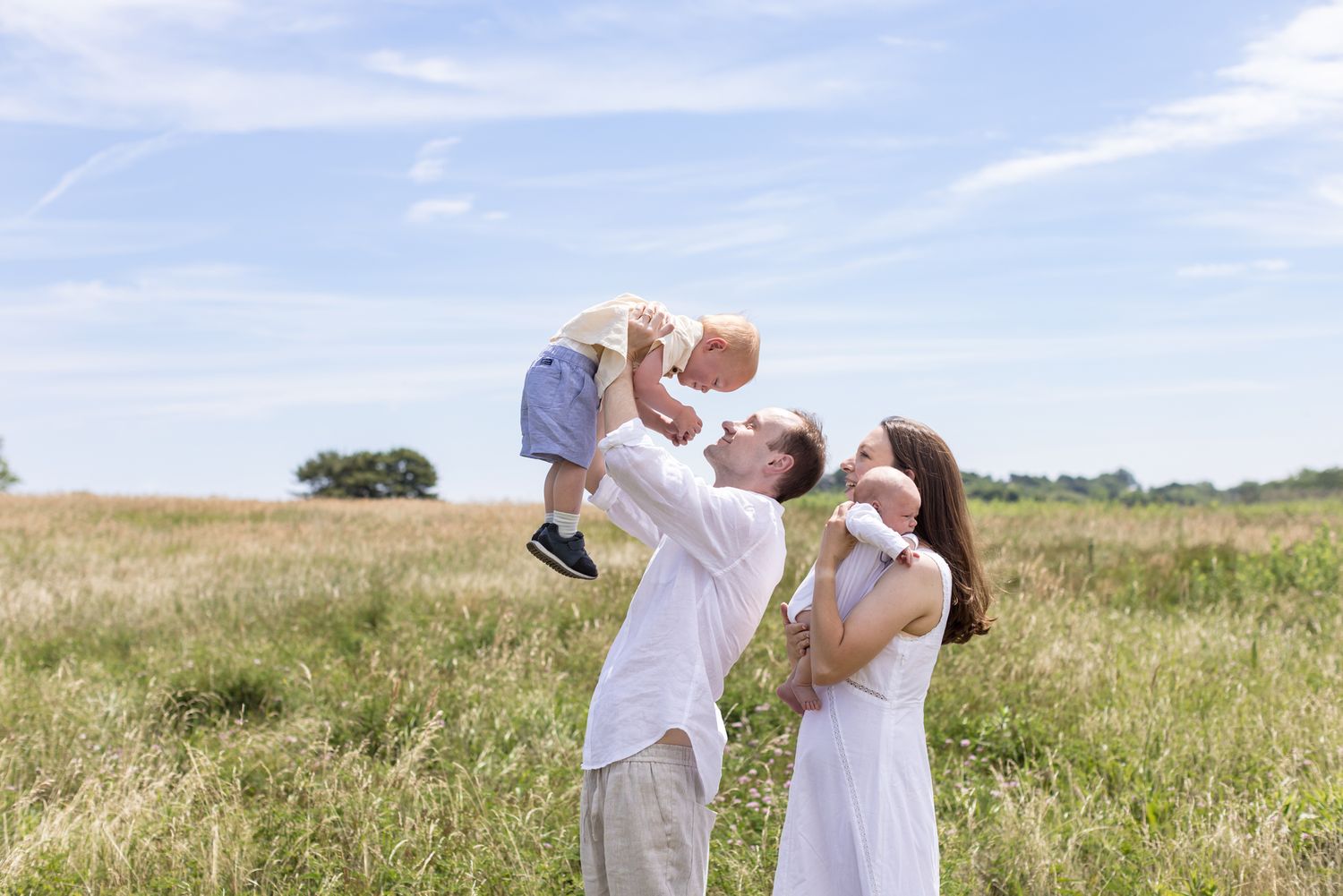 A father holds his toddler son up in the air as wife looks on holding newborn son over her shoulder in a grassy field on a sunny day.