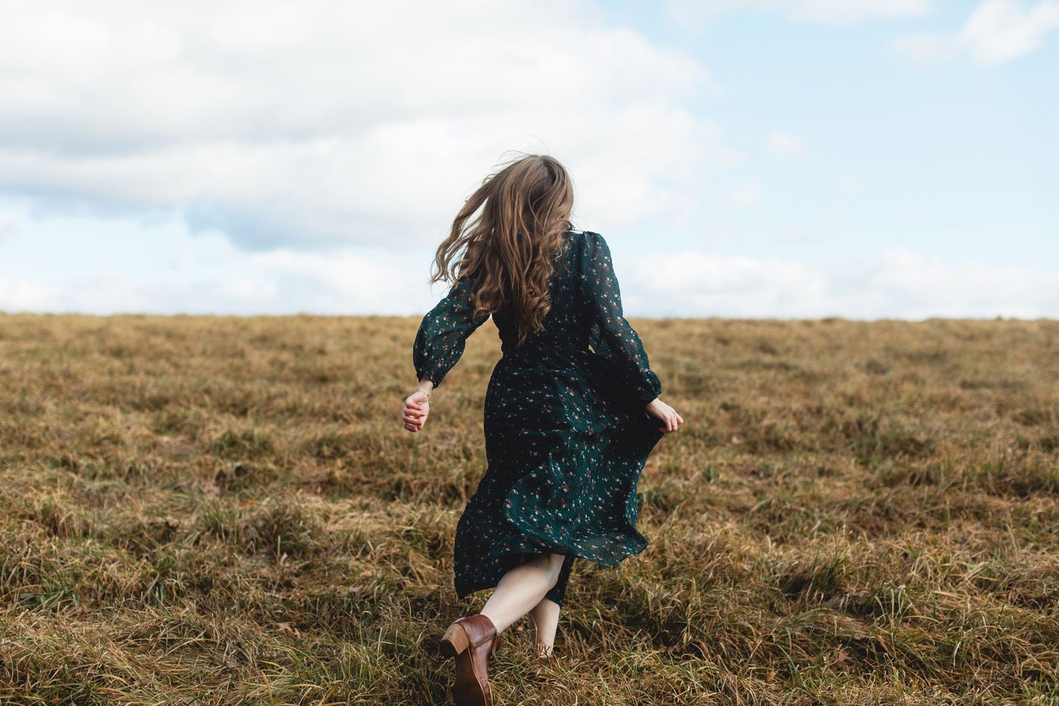 A lady in a dark teal dress runs through a golden autumn field on a cloudy day for a portrait session at Ward Hill in North Andover, MA.