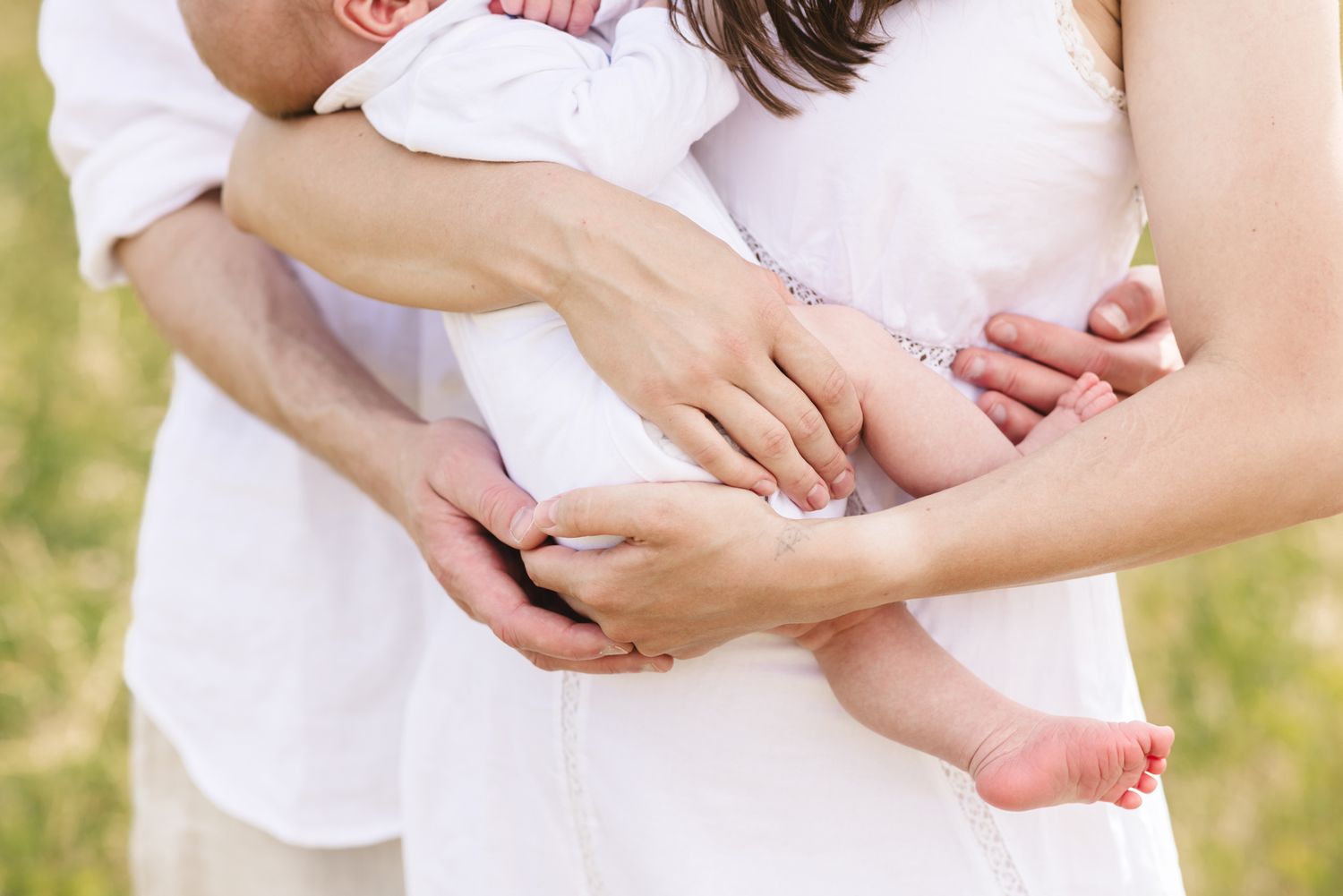 A cropped photo of a couple wearing white embracing their newborn for newborn photos with grass in the background in Winthrop.