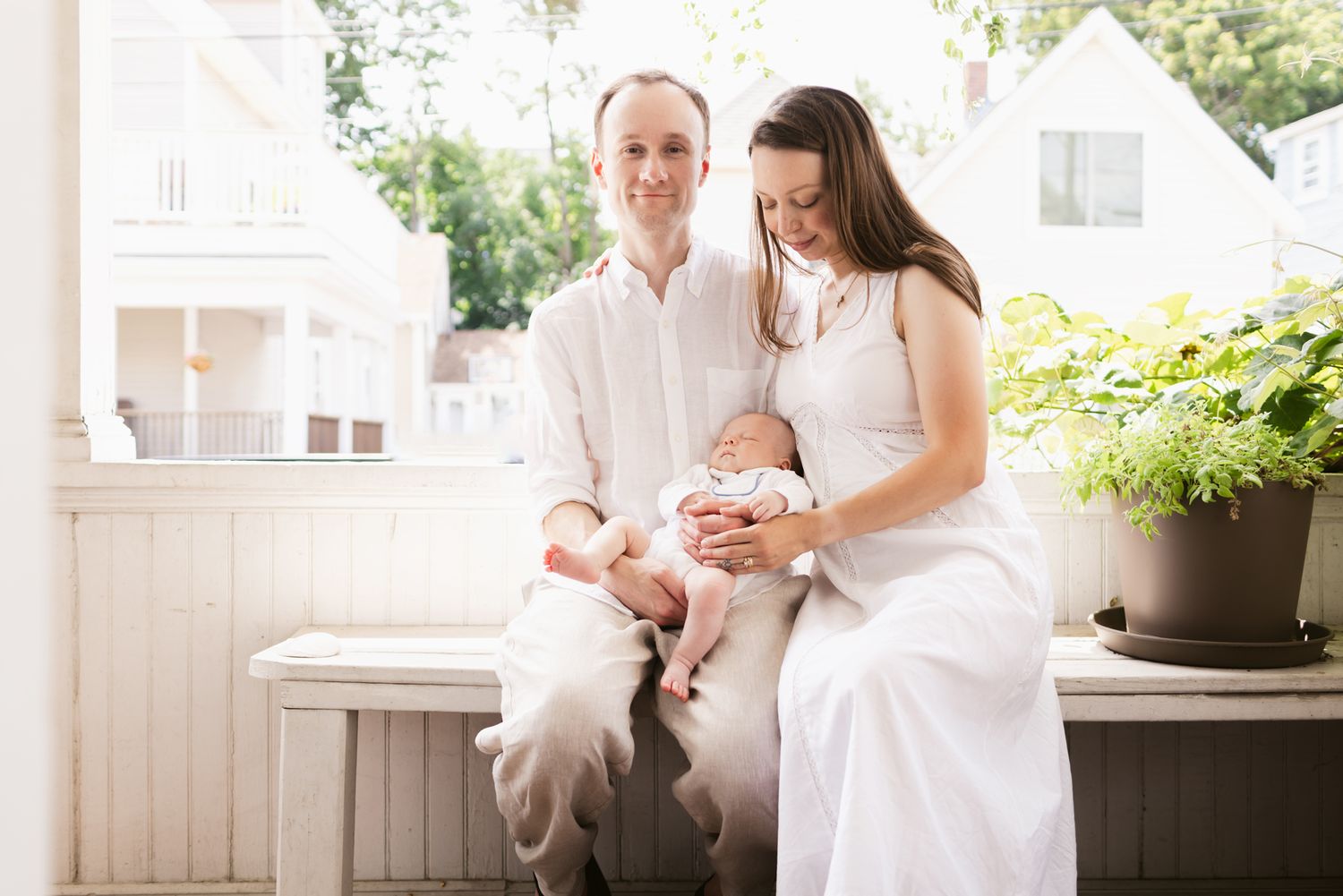 Father and mother wearing white sit on a porch on a white bench on a sunny day holding their newborn on their lap for newborn photos.