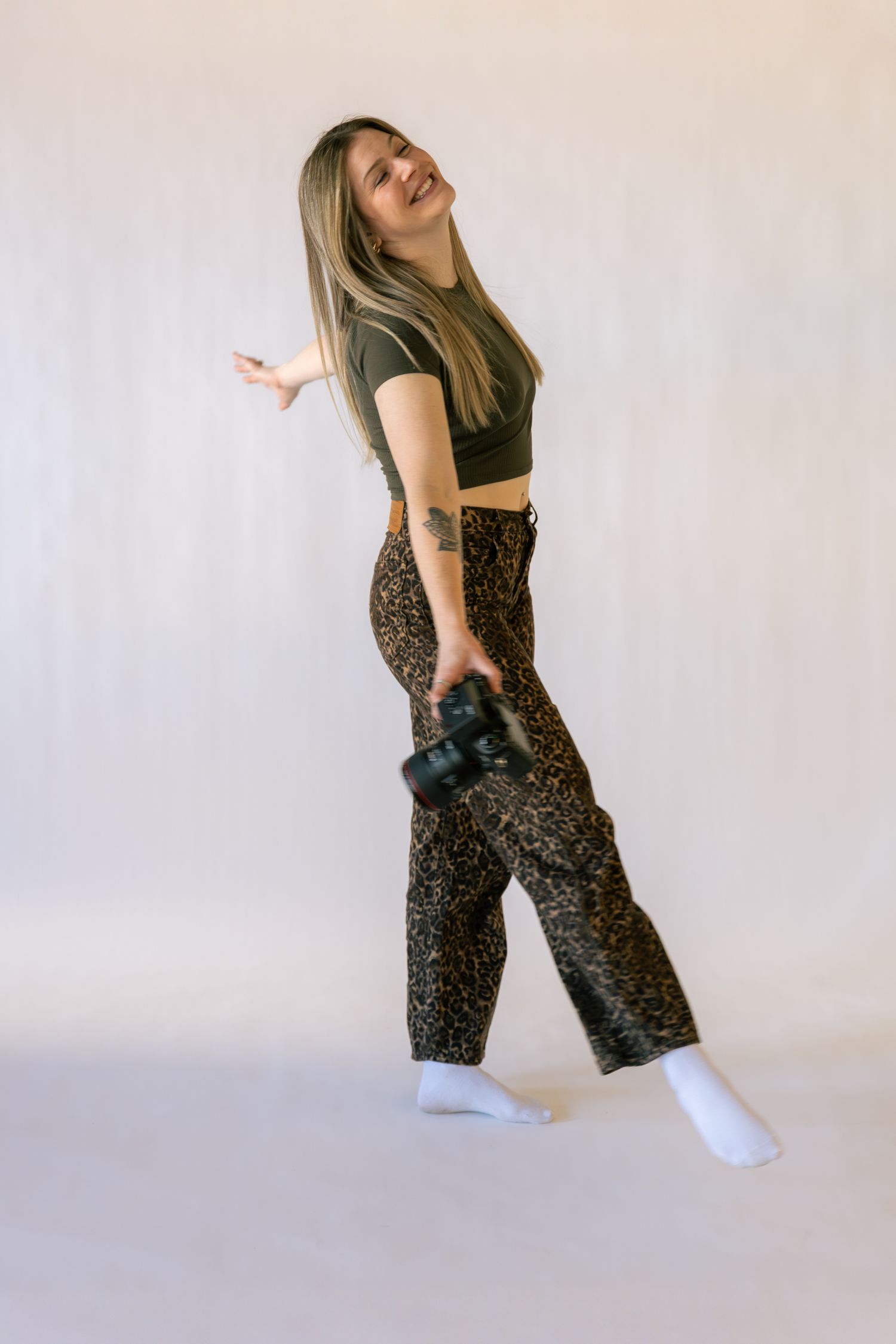 Young woman in crop top and leopard print pants holds a camera, smiling and posing against a white background.