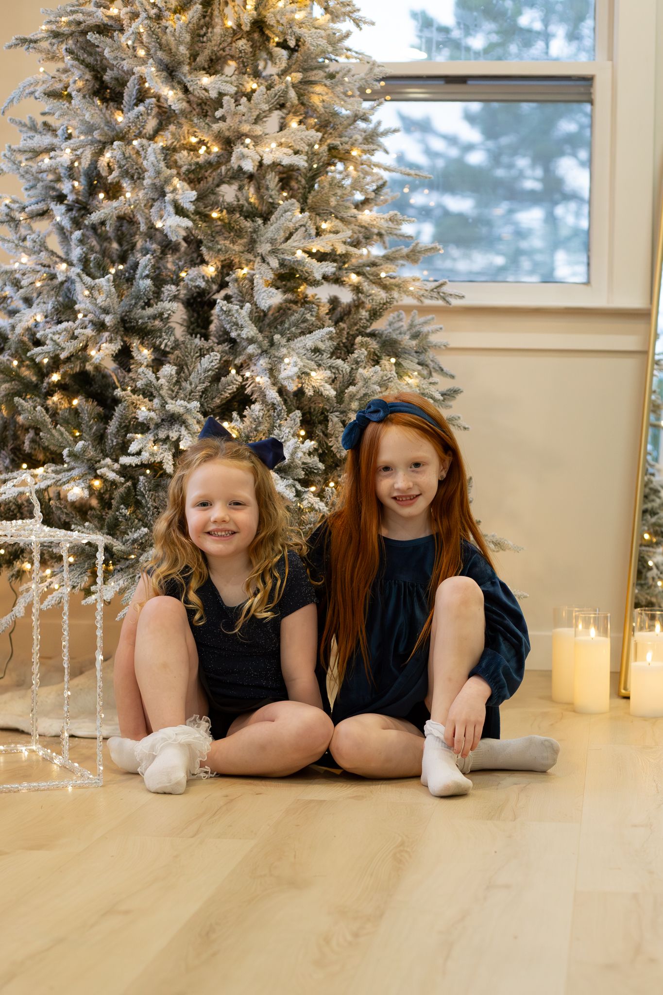 Two young girls in navy dresses sitting by decorated flocked Christmas tree with candles.