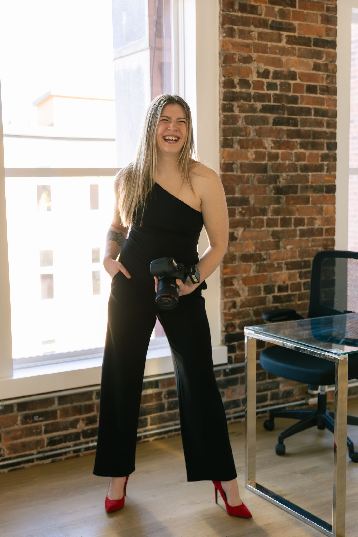 Woman in black jumpsuit and red heels holding a camera, smiling in a bright loft studio with brick wall.