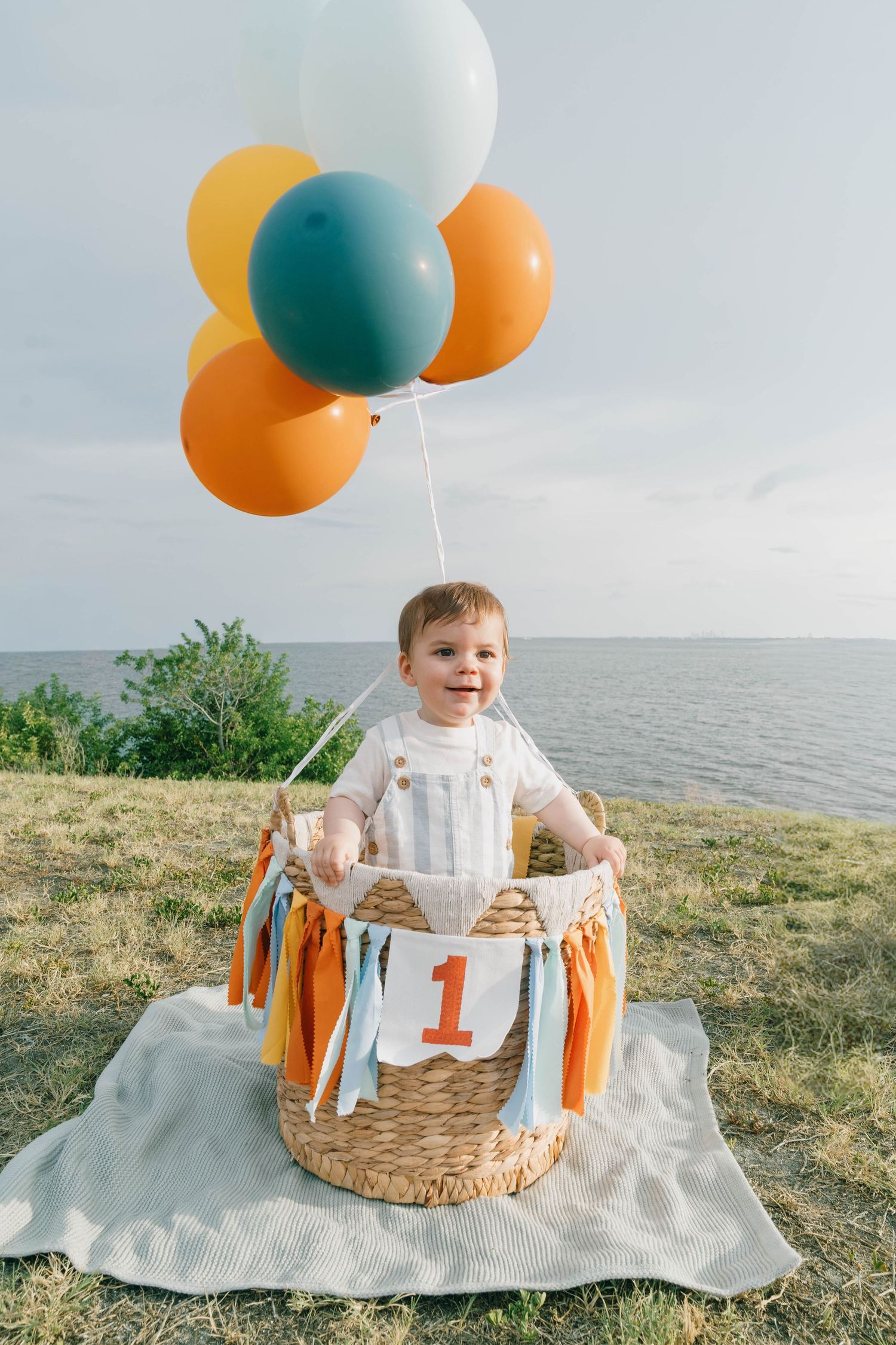 A smiling baby sits in a wicker basket decorated with colorful balloons and a number 1 sign, celebrating their first birthday outdoors.
