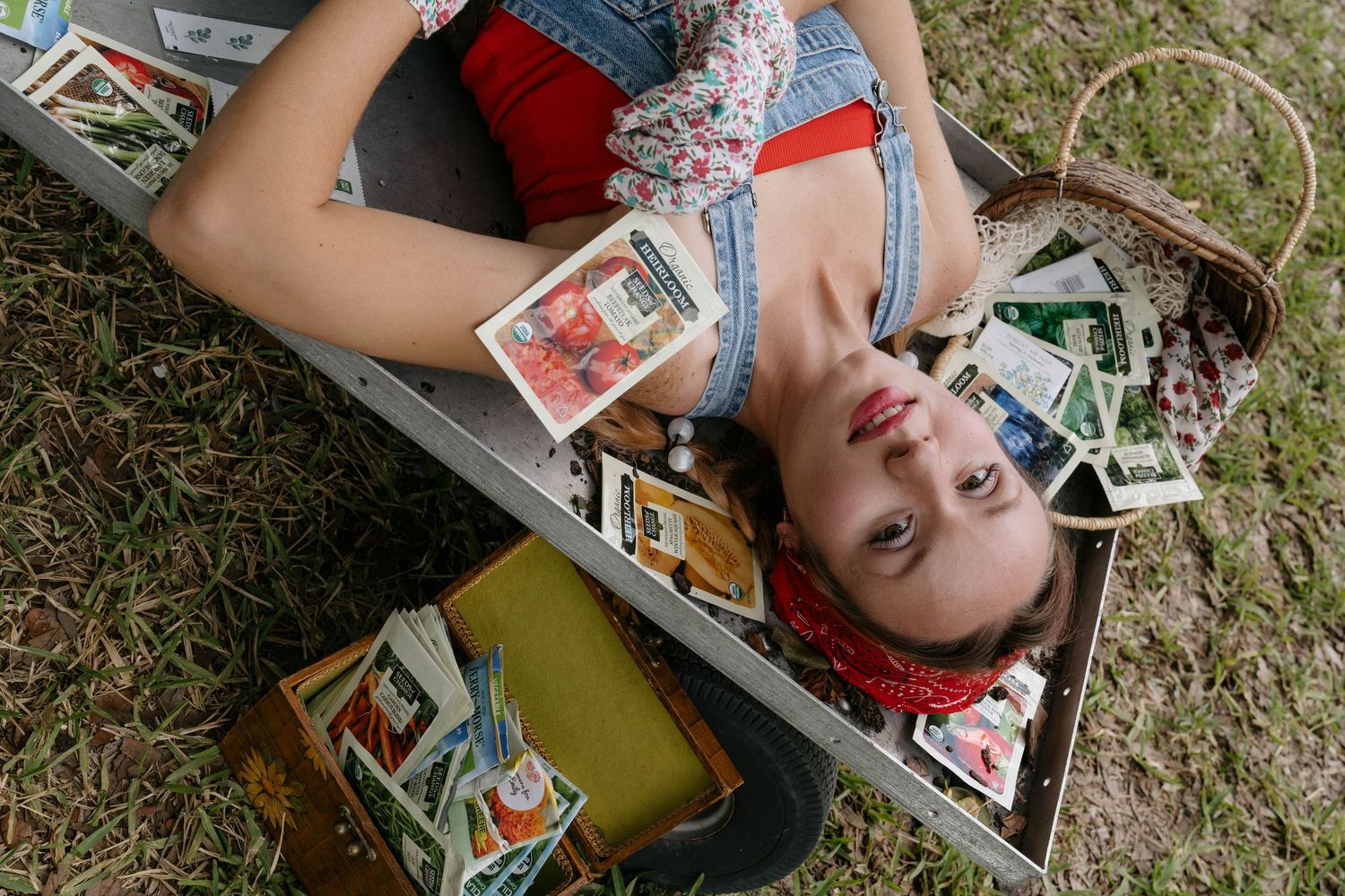 Young woman lying in a vintage suitcase surrounded by colorful postage stamps and retro collectibles outdoors.