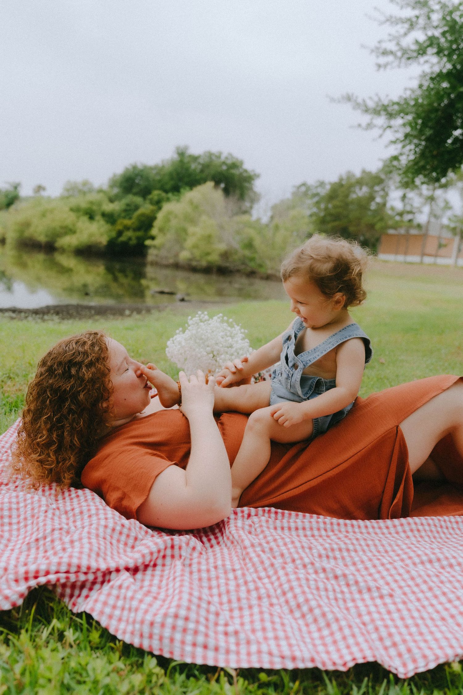 An outdoor picnic scene with people bonding and playing on a red checkered blanket near a pond.