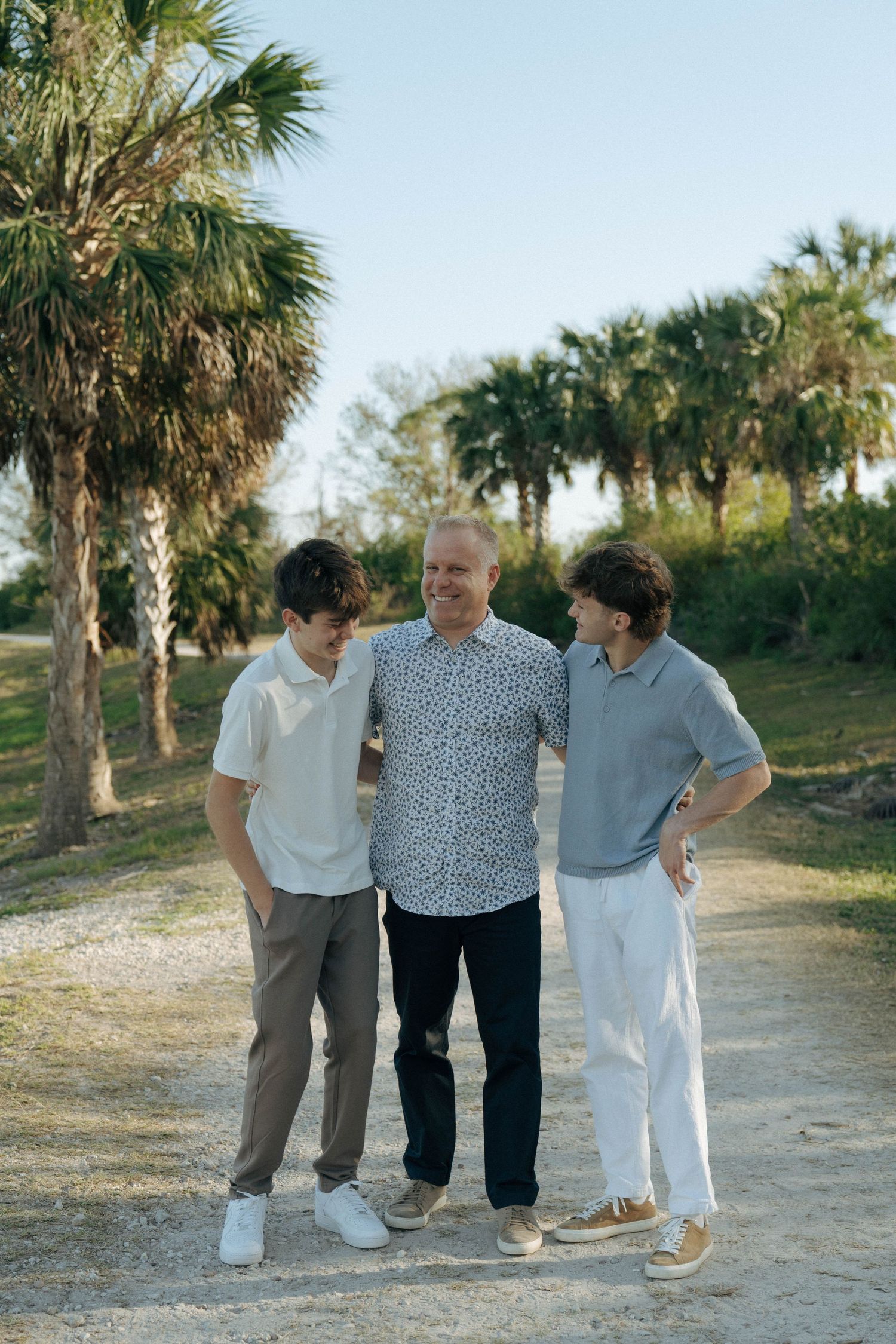 Three people in casual attire standing together on a sandy path lined with palm trees in a tropical setting.