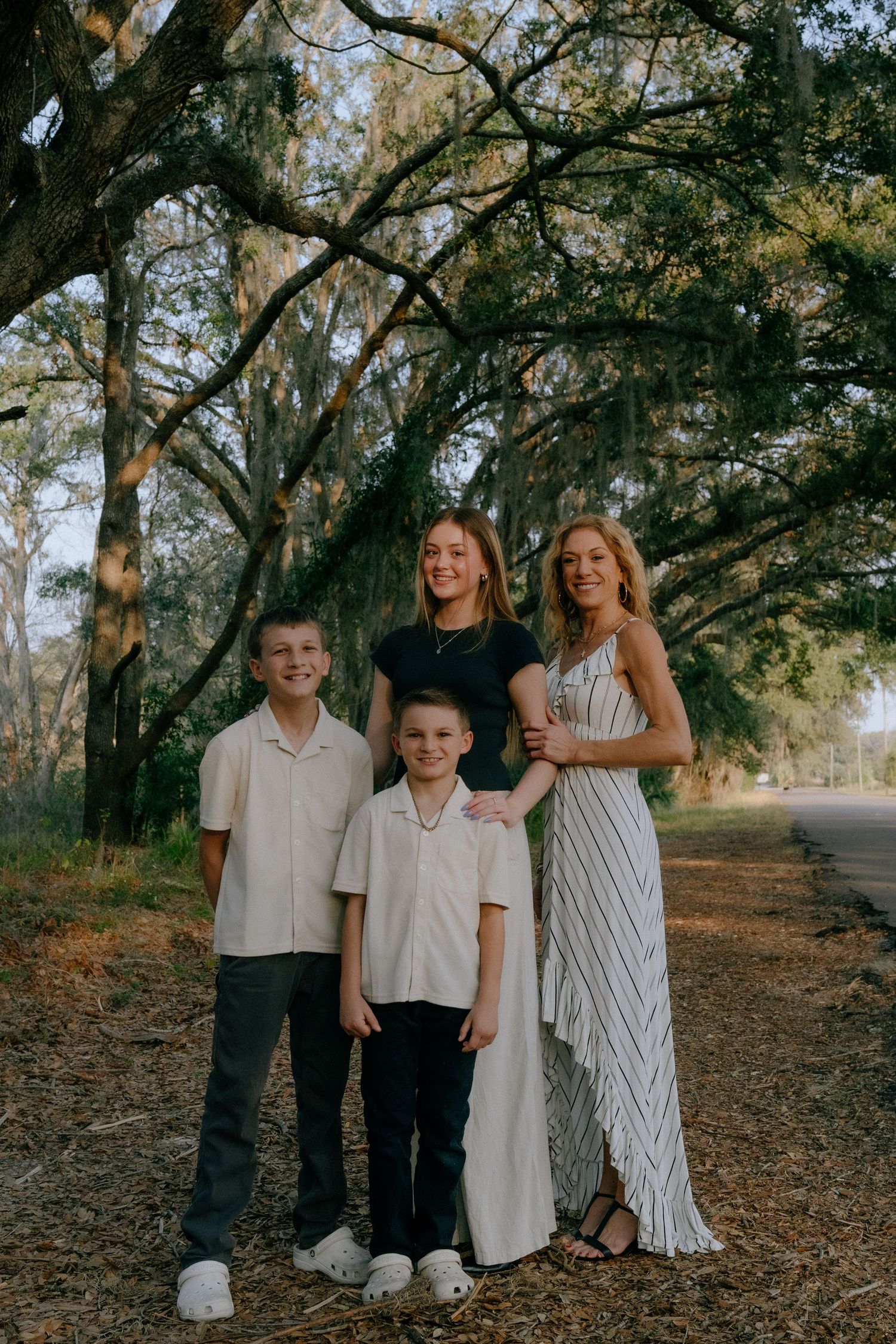 A family poses together outdoors on a path lined with Spanish moss-draped trees in natural lighting.