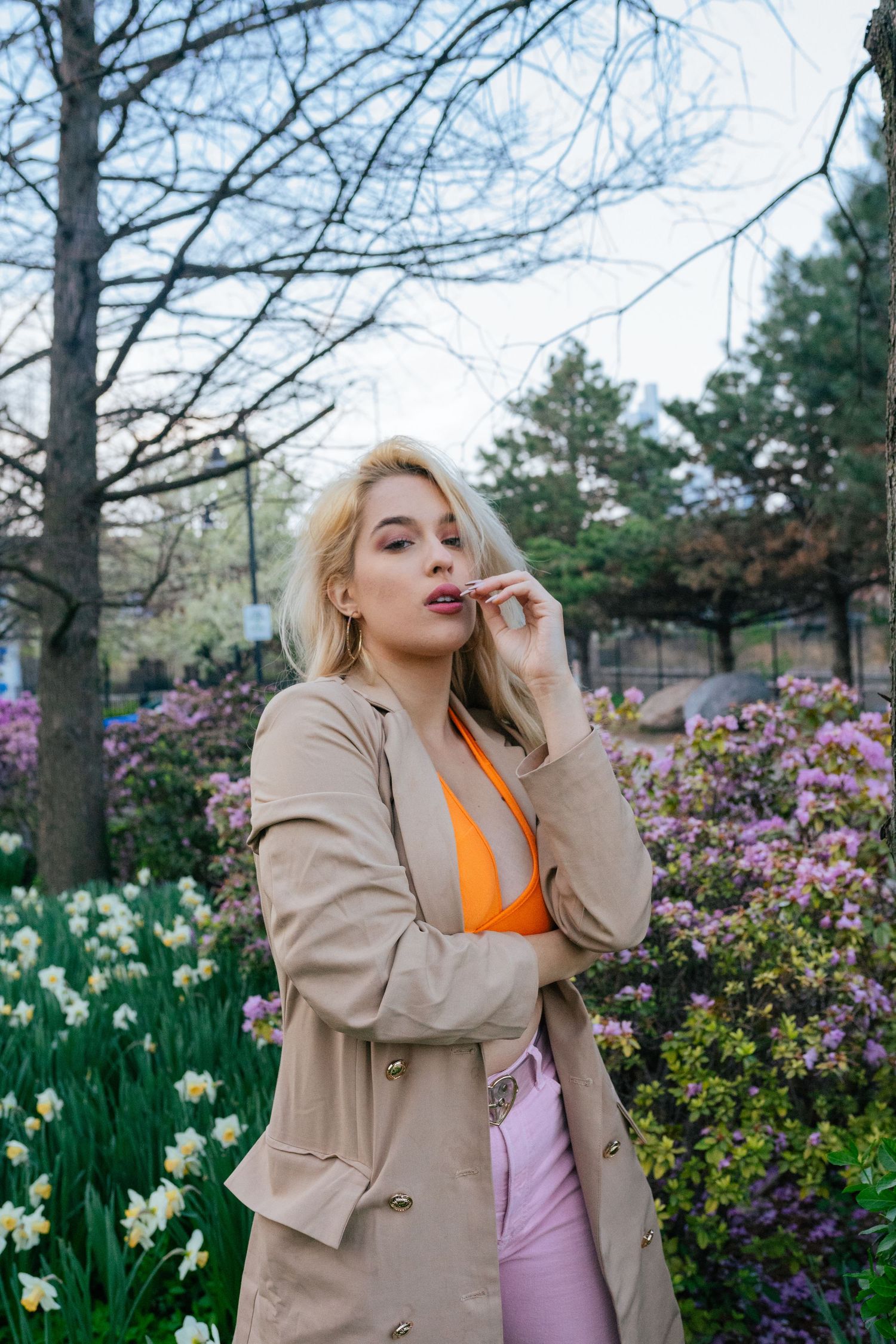 Woman in beige coat and orange top poses in spring garden with blooming flowers and white daffodils.