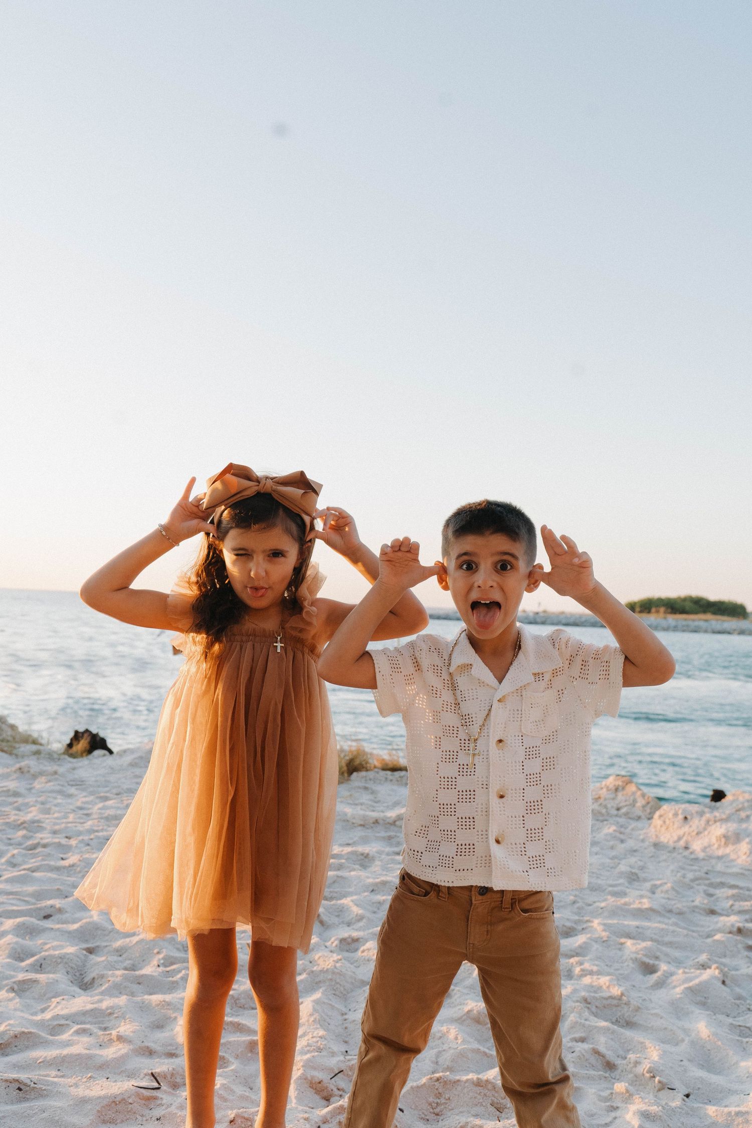 Two children making funny faces on a sandy beach at sunset, girl in tan dress and boy in white shirt.
