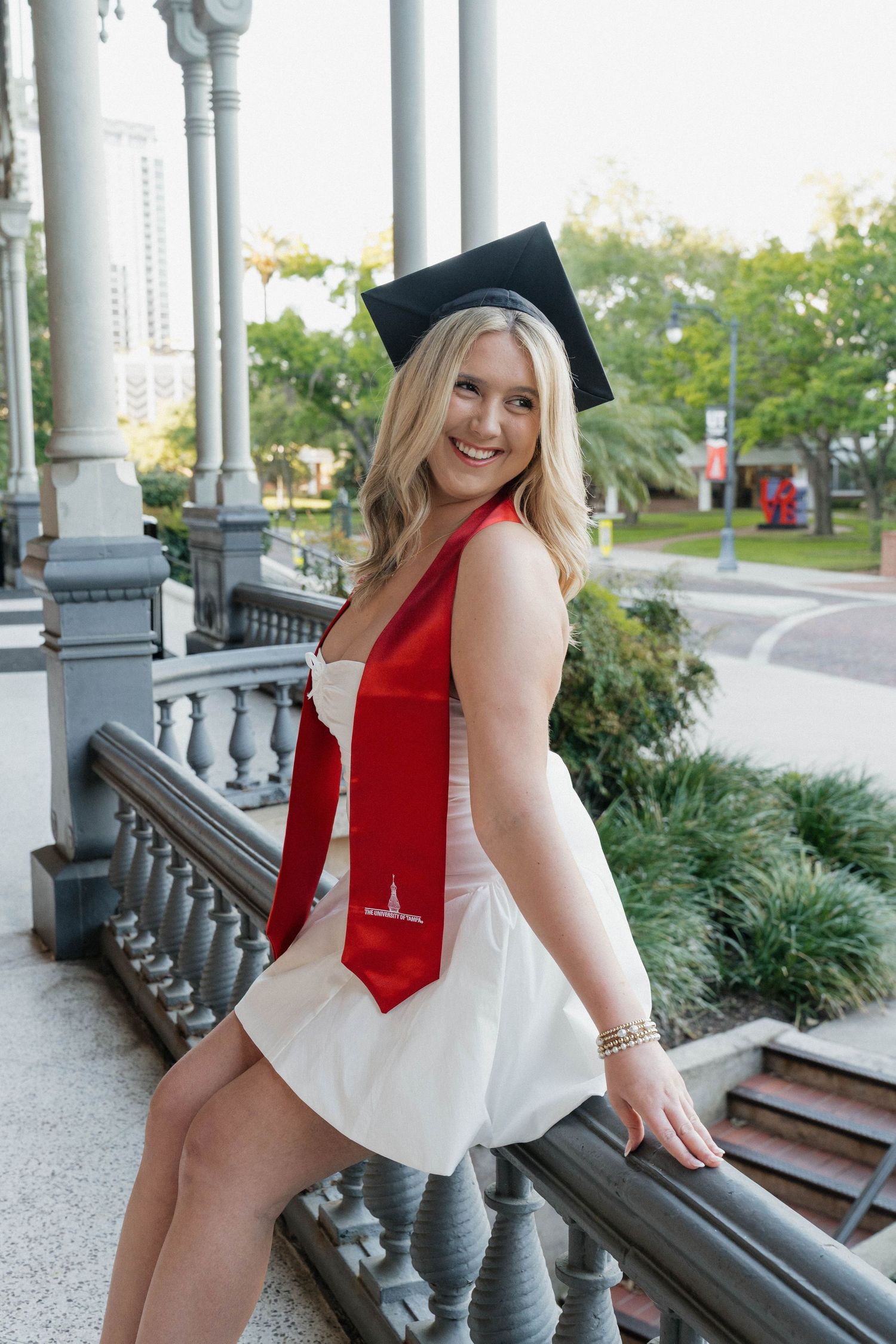 Smiling blonde graduate wearing cap and red stole with white dress poses on ornate stone staircase outdoors.