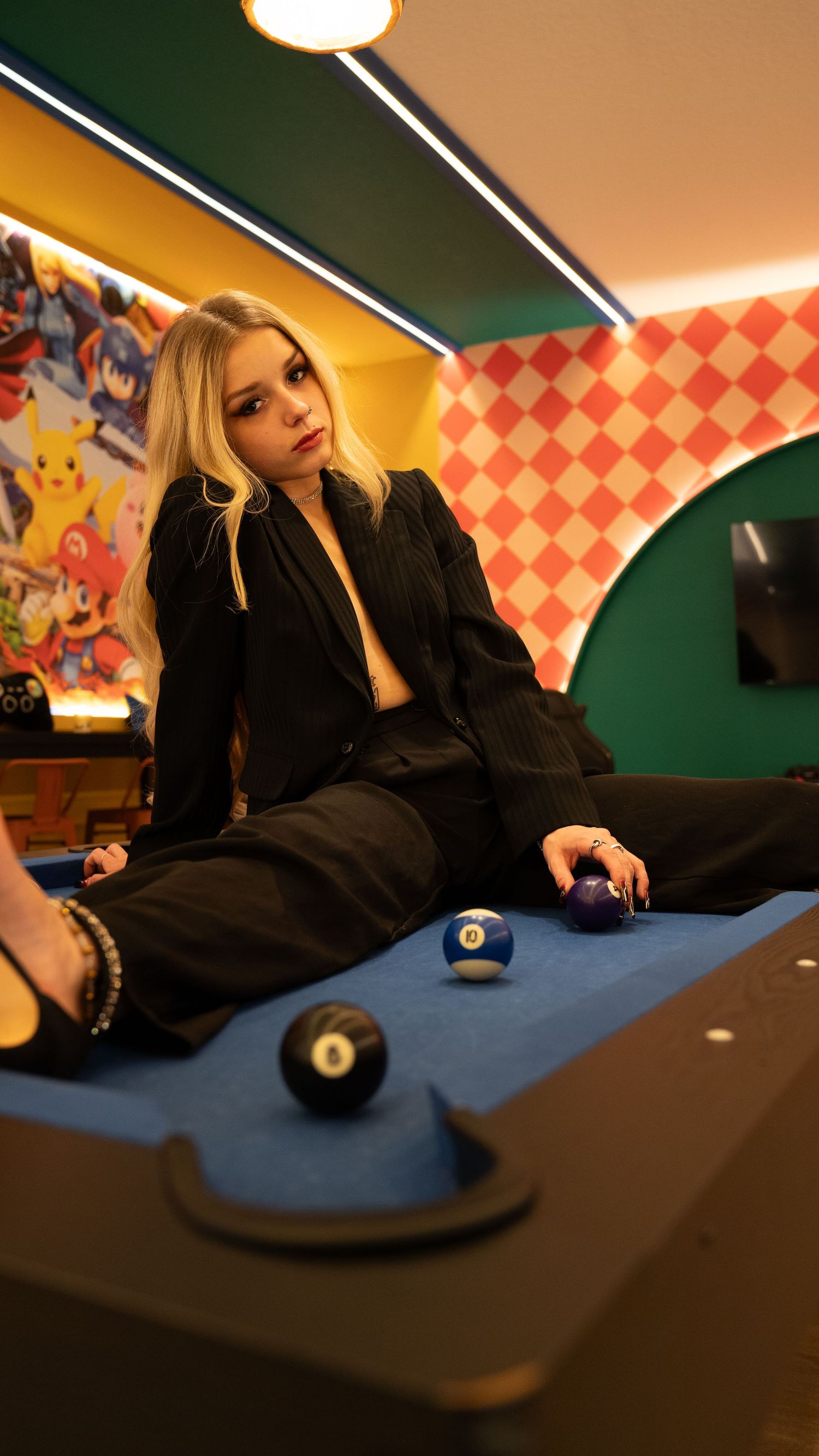 A stylish figure in black attire poses on a pool table in a retro-themed gaming room with checkered walls and moody lighting.